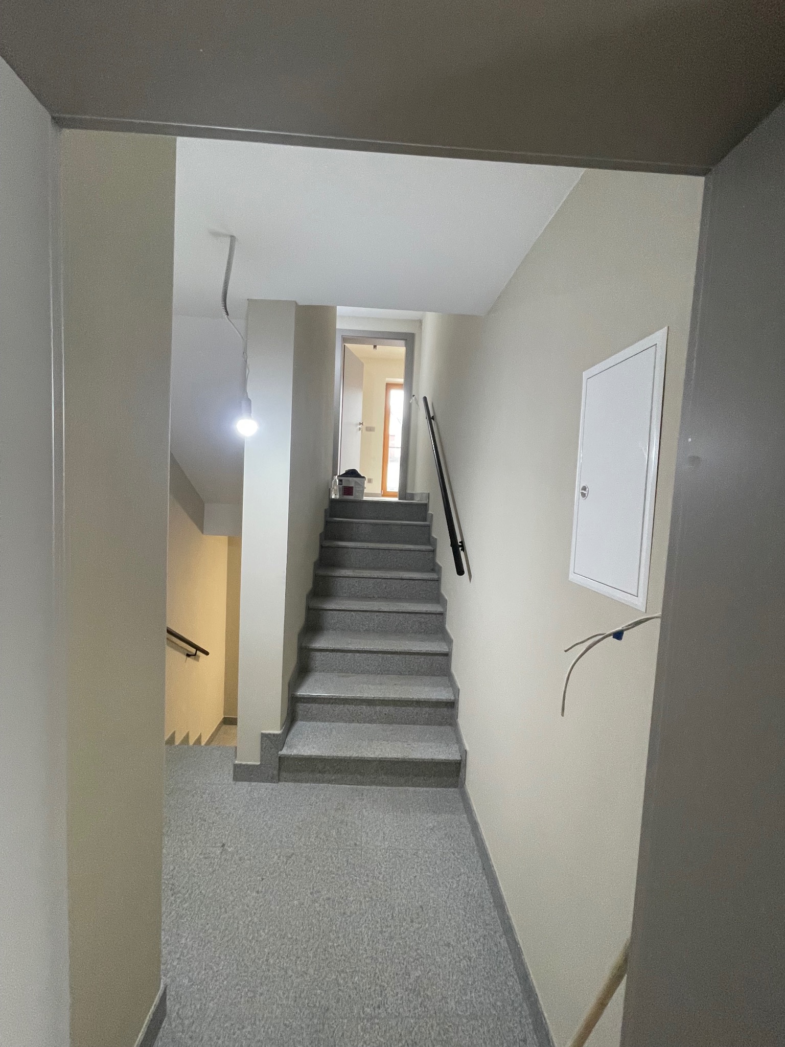 Klatka schodowa w budynku wielorodzinnym z szarymi schodami i beżowymi ścianami. Widoczne piony instalacyjne i oświetlenie. Ujęcie z dołu schodów.