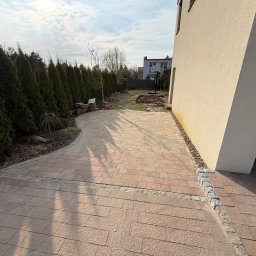 JANBUD Specjalistyczne Przedsiębiorstwo Budowlane Janusz Nowaczyk - Ułożona, beżowo-czerwona kostka brukowa tworząca ścieżkę w ogrodzie, obok domu z jasną elewacją i rzędem wysokich, zielonych tuj. Widok na ogród i fragment budynku.