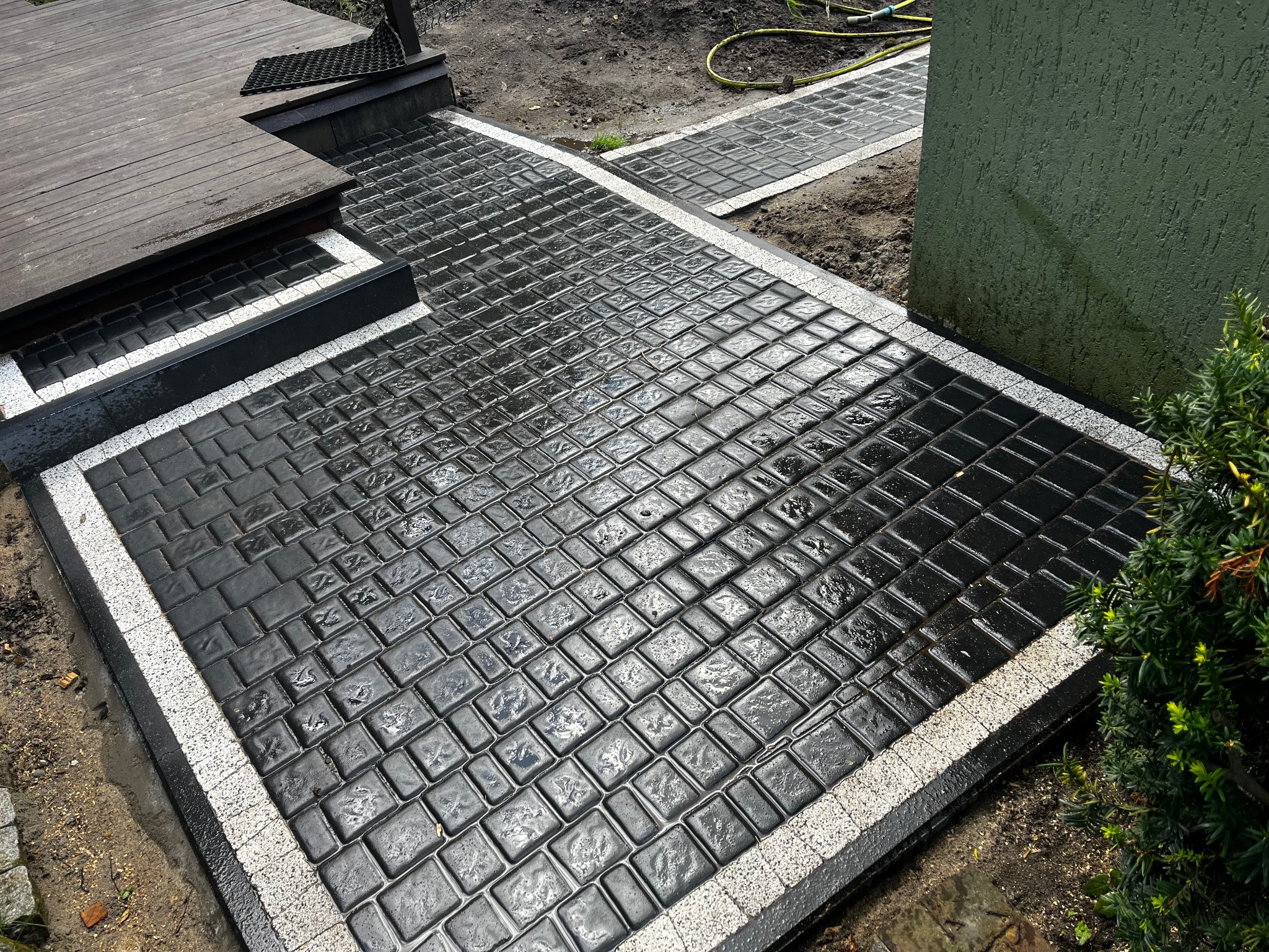 Ułożona, czarna kostka brukowa z jasną obwódką, prowadząca od drewnianego tarasu. Widoczny fragment trawnika i zielona ściana budynku. Kostka mokra, prawdopodobnie po deszczu.