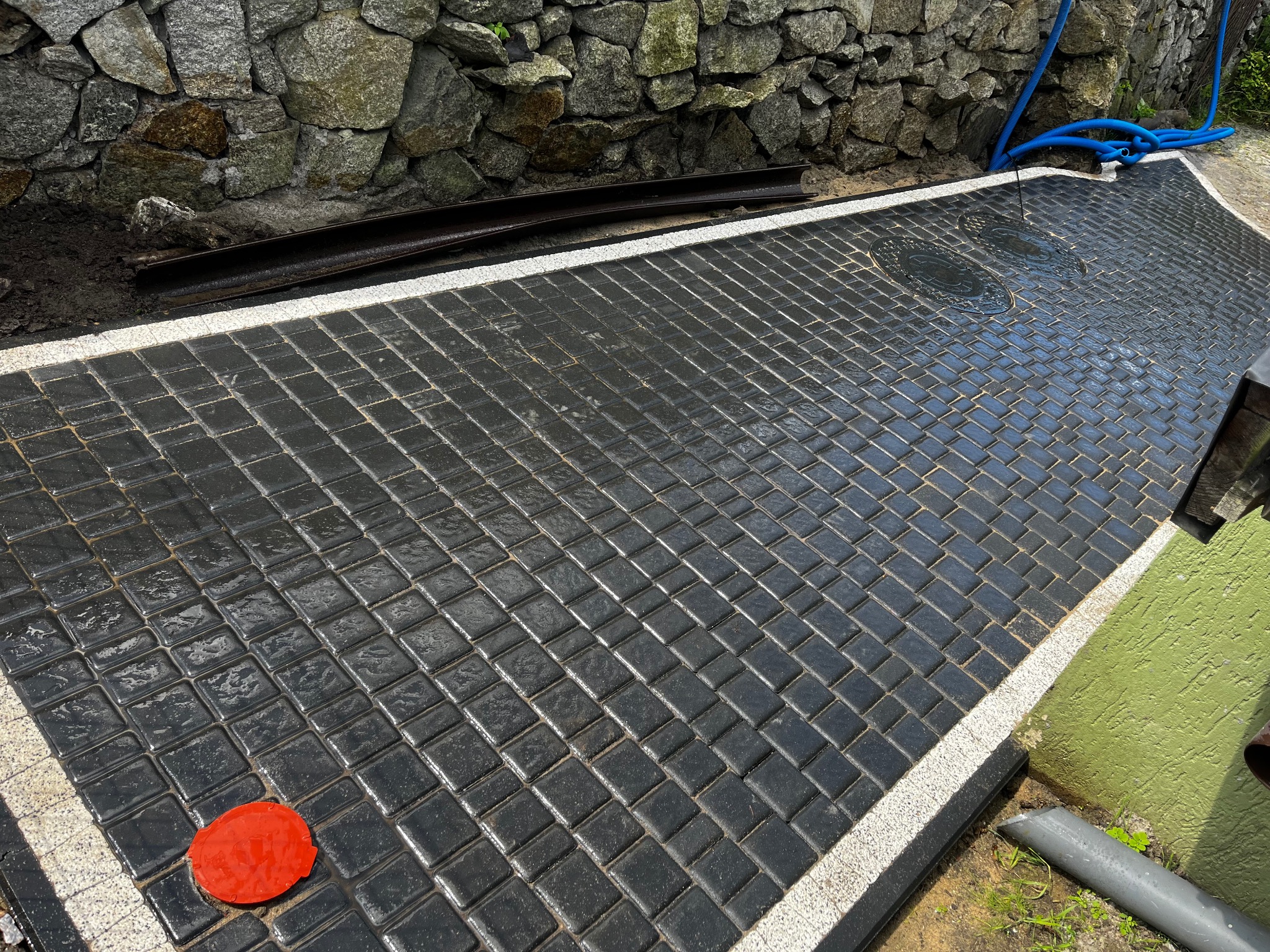 Ułożona, czarna kostka brukowa z białym obramowaniem, widoczne studzienki kanalizacyjne i czerwony element na powierzchni. W tle kamienny murek i niebieski wąż.