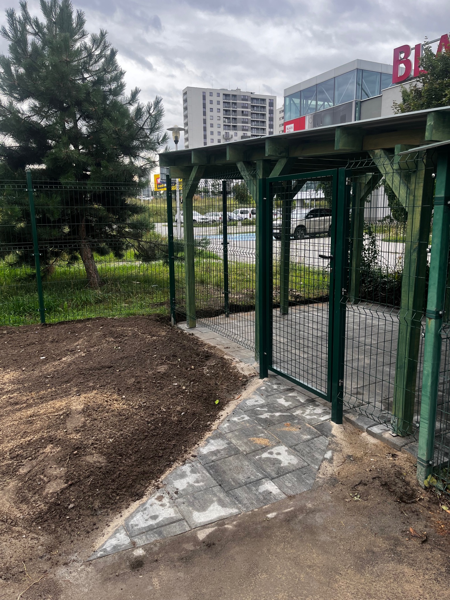 Zielony, metalowy płot z furtką i zadaszeniem, obok świeżo przekopana ziemia i ułożona kostka brukowa. W tle budynki miejskie i drzewo iglaste.