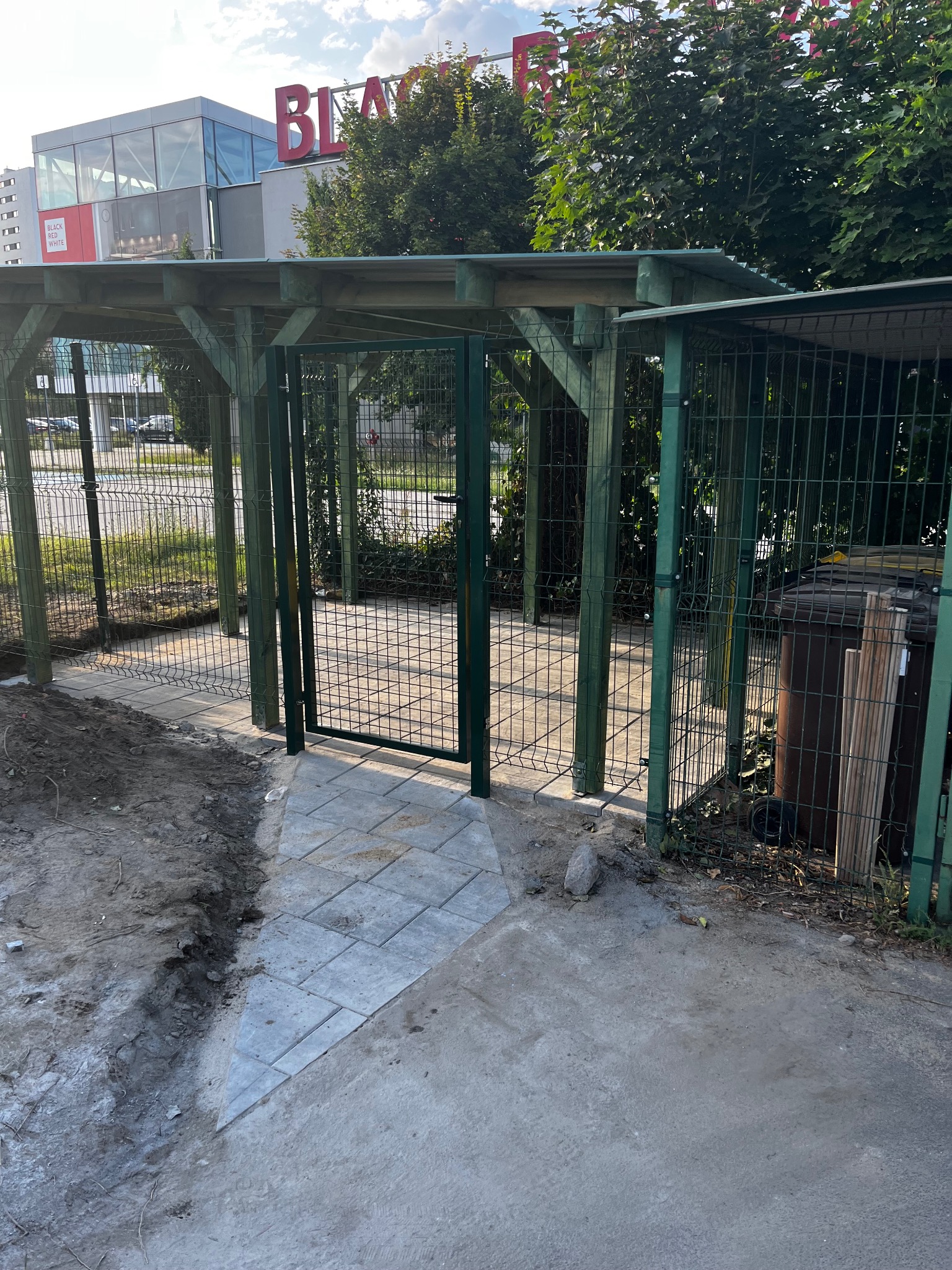 Zadaszona wiata śmietnikowa z zielonej siatki i drewnianych słupów, z otwartą furtką i fragmentem chodnika z szarej kostki. W tle budynek z szyldem.