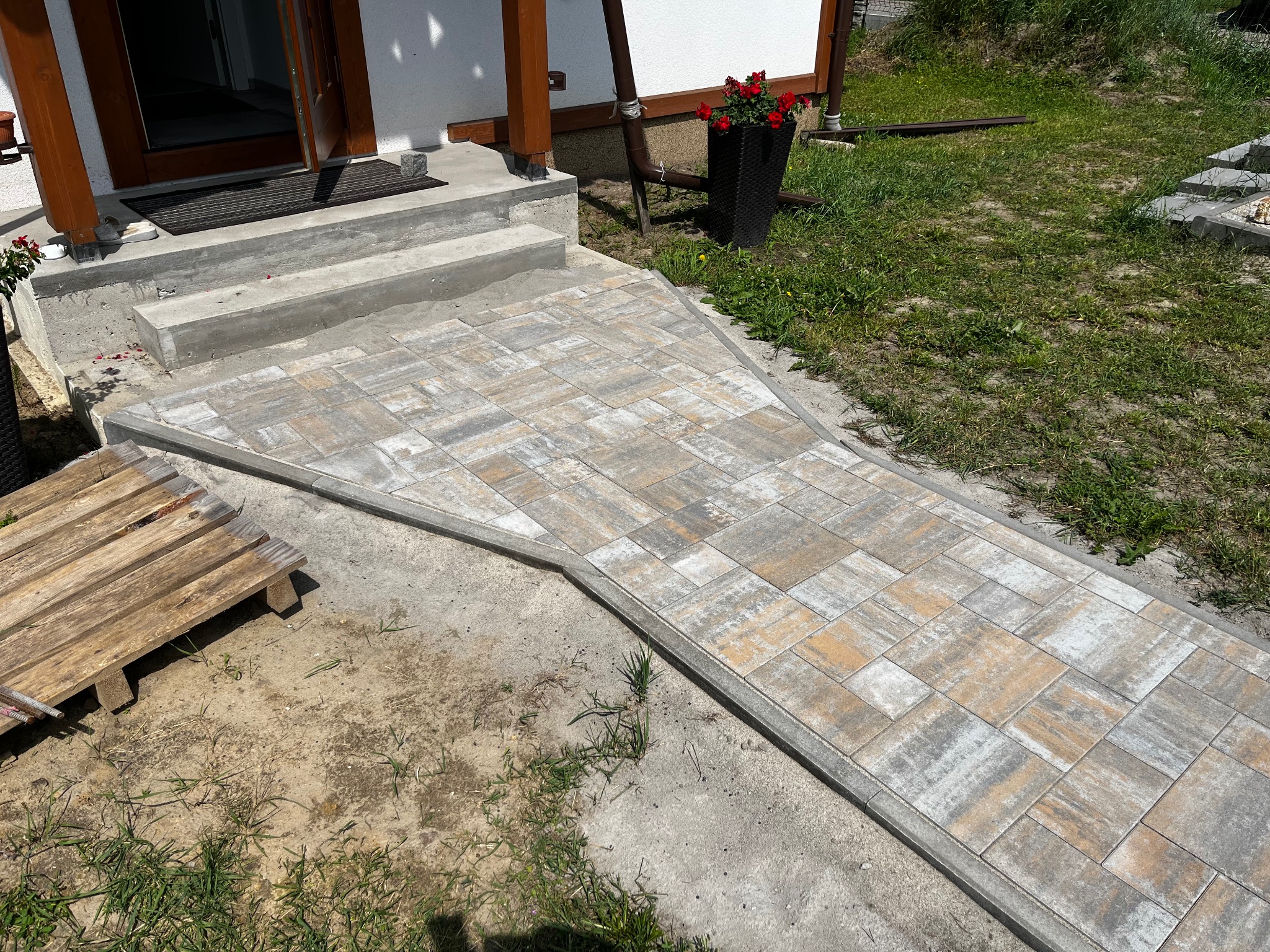 Ułożona ścieżka z kostki brukowej prowadząca do wejścia domu, z widocznymi schodami i elementami zieleni w tle. Paleta drewniana leży obok ścieżki.