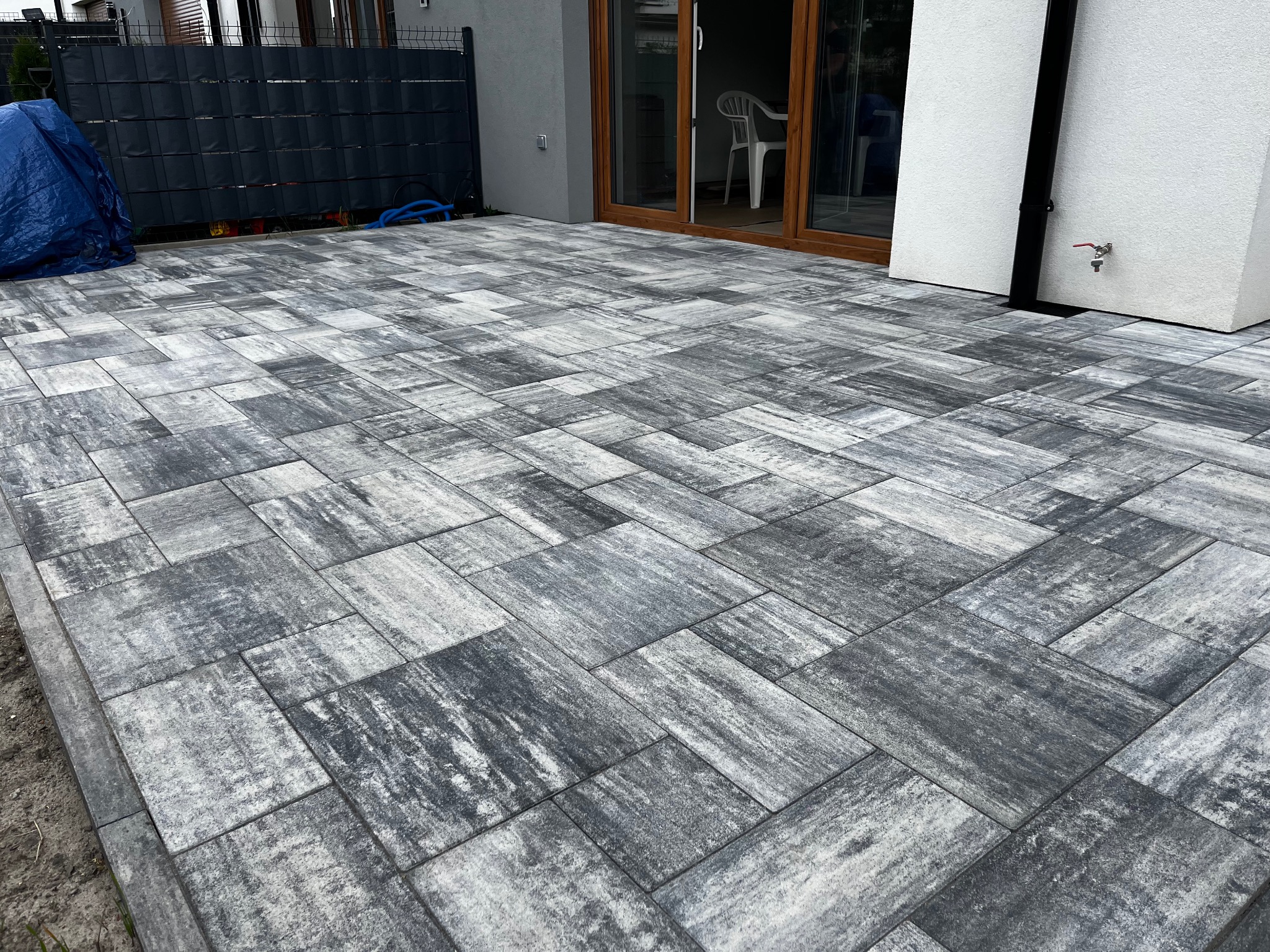 Ułożona szara kostka brukowa na posesji, widok z góry. W tle czarne ogrodzenie panelowe i fragment budynku z oknami. Kostka ułożona w nieregularny wzór.