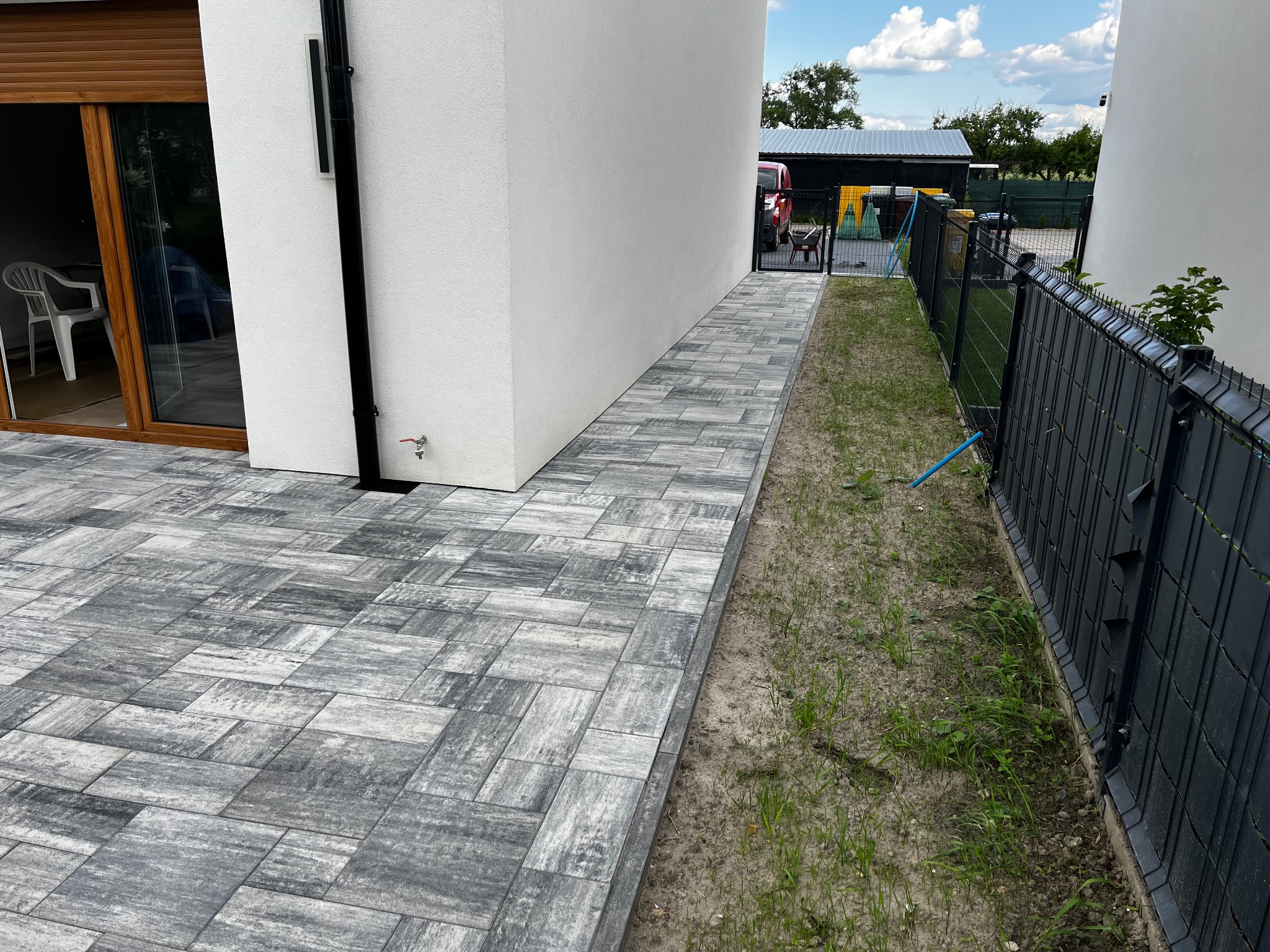 Ułożona szara kostka brukowa prowadząca wzdłuż białego budynku, z czarnym ogrodzeniem po prawej stronie. Widoczny fragment ogrodu i błękitne niebo.