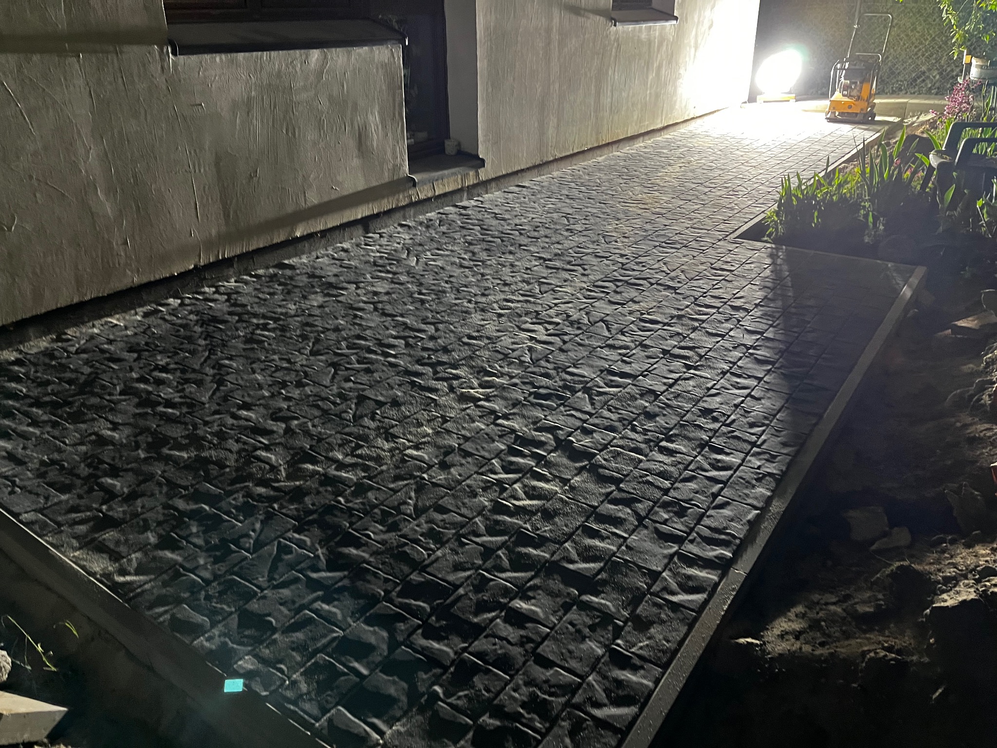 Ułożona, ciemna kostka brukowa wzdłuż budynku, oświetlona lampą budowlaną. Widoczna ubijarka. Krawężniki wyznaczają obszar robót.