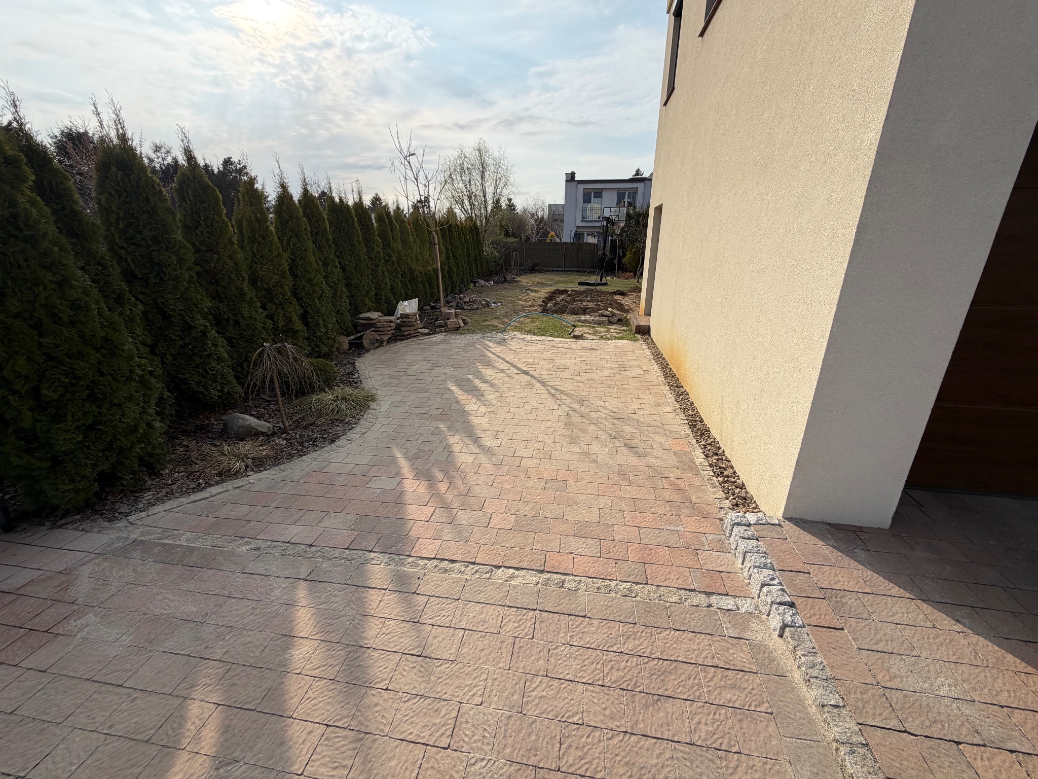 Ułożona, beżowo-czerwona kostka brukowa tworząca ścieżkę w ogrodzie, obok domu z jasną elewacją i rzędem wysokich, zielonych tuj. Widok na ogród i fragment budynku.