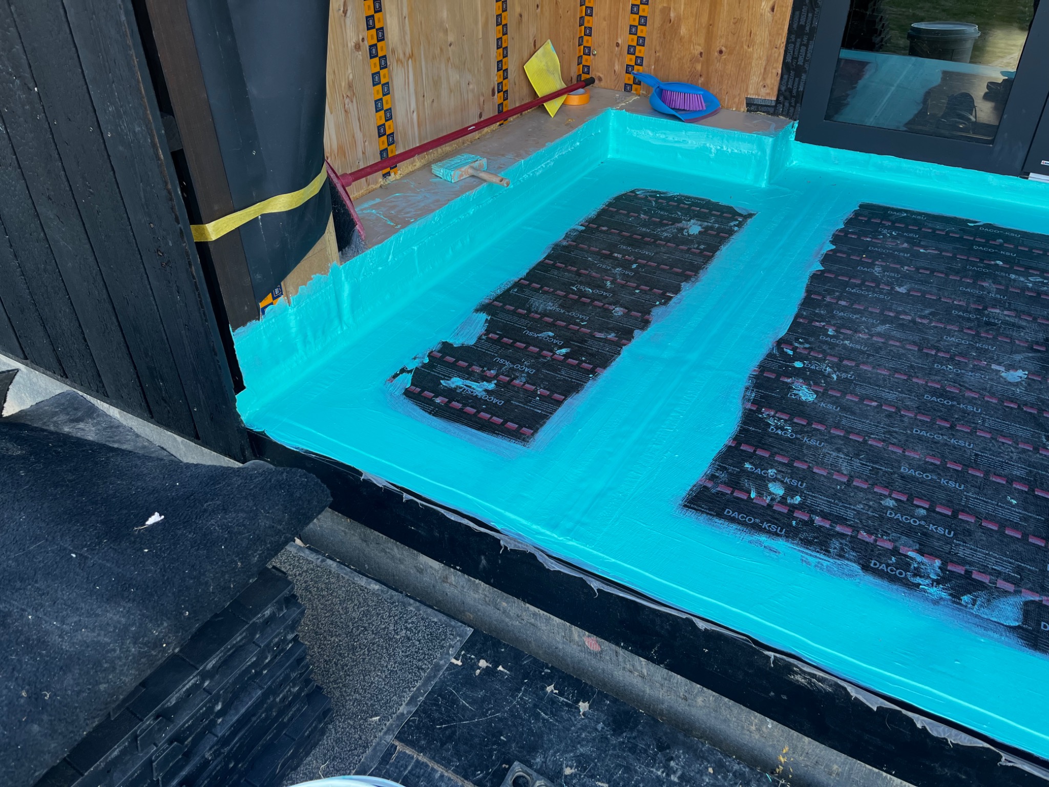 Hydroizolacja balkonu w trakcie wykonywania, widoczne pasy izolacyjne DACO KSU, świeżo nałożona turkusowa warstwa hydroizolacyjna, pędzel i miotła w tle.
