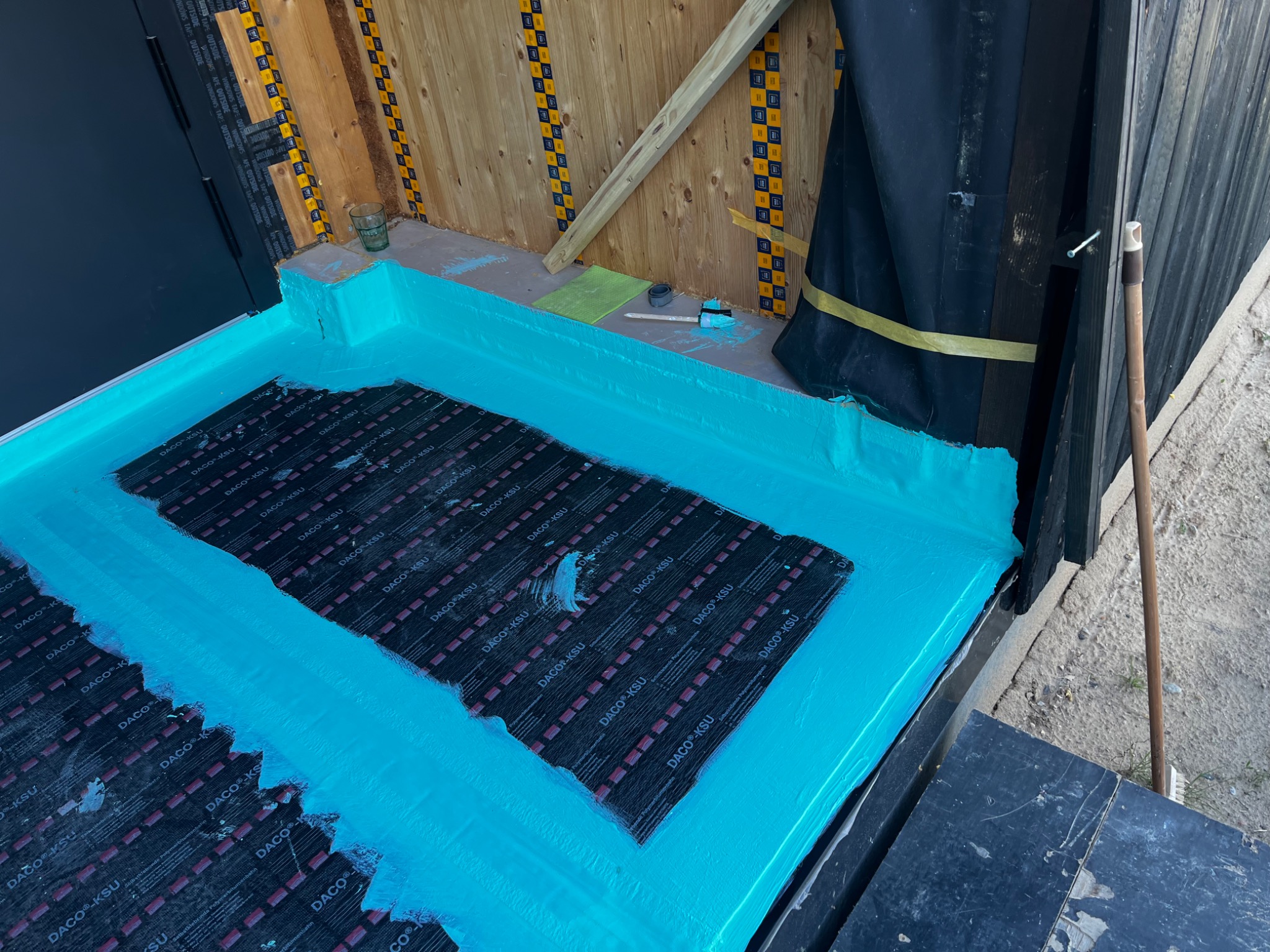 Hydroizolacja płynną membraną w kolorze turkusowym na dachu domu modułowego, widoczne arkusze papy podkładowej DACO KSU i narzędzia pracy.