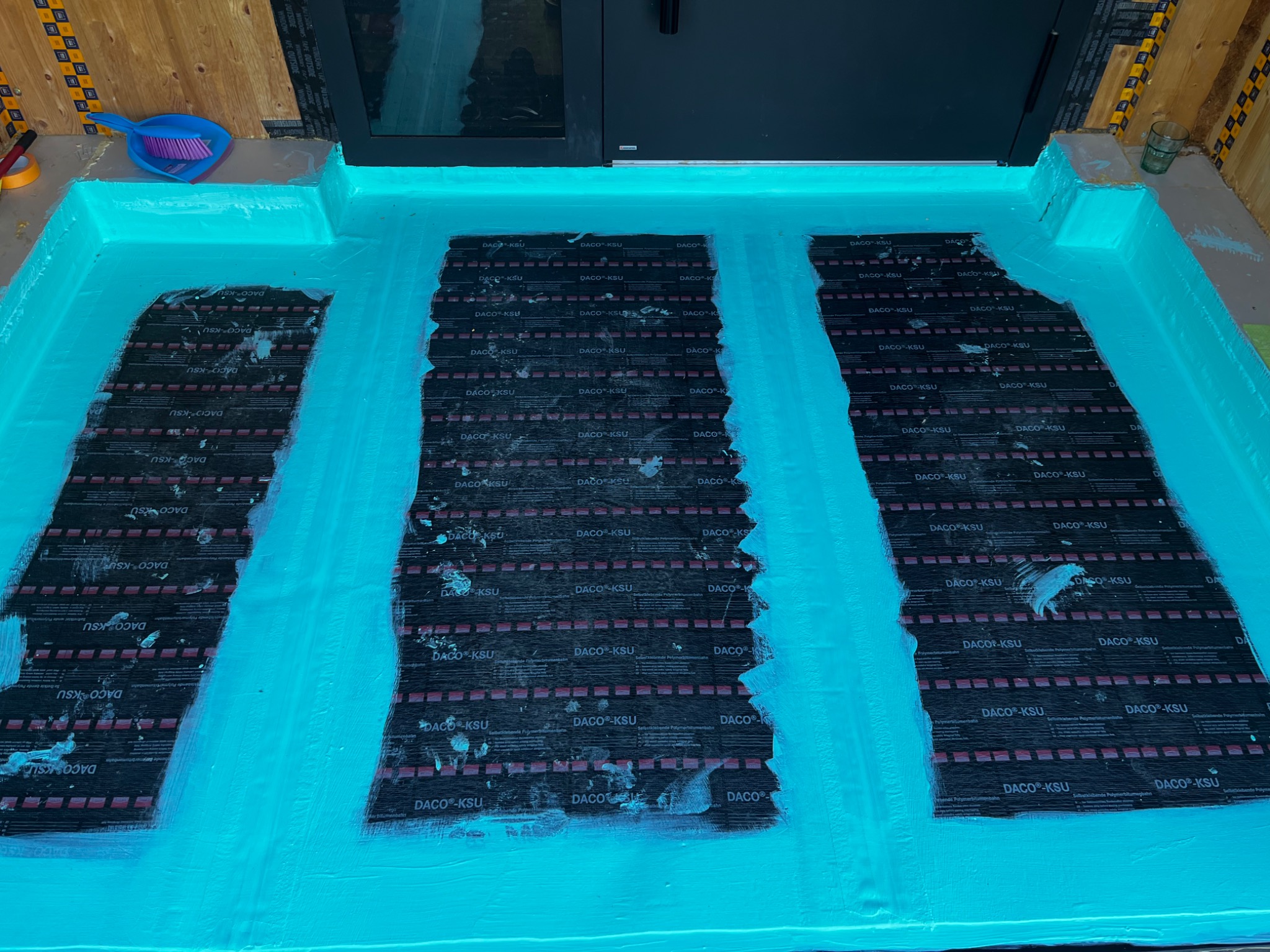 Hydroizolacja podłoża wewnątrz budynku, pokryta warstwą uszczelniającą w kolorze turkusowym, z widocznymi fragmentami czarnej membrany izolacyjnej Daco-KSU.