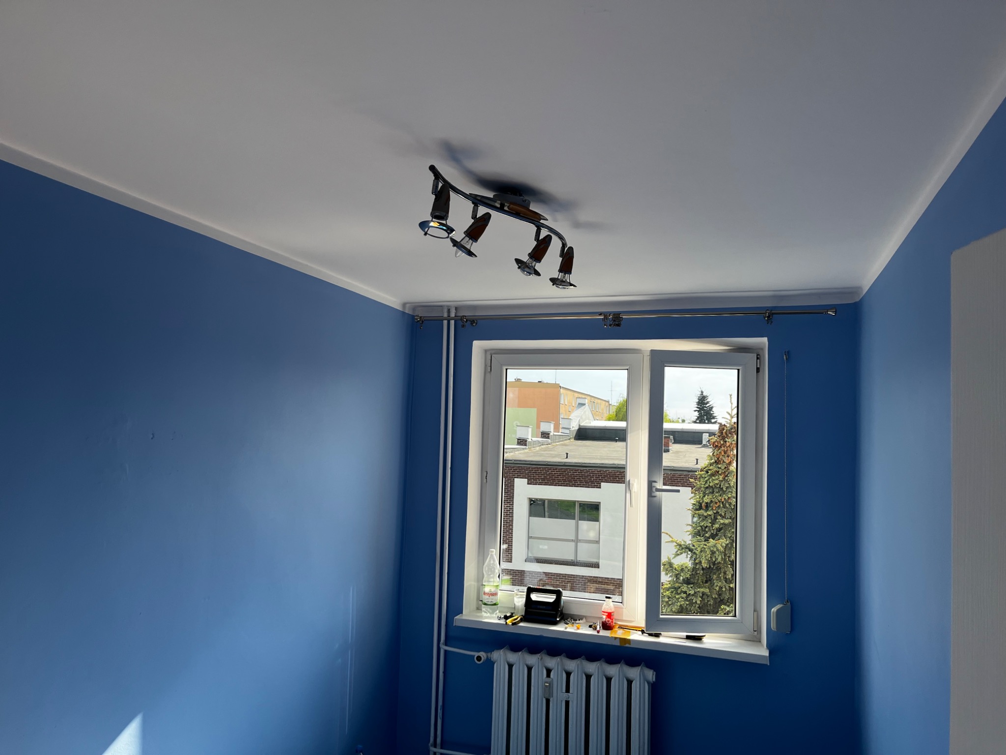 Świeżo pomalowany pokój z niebieskimi ścianami, białym sufitem i oknem z widokiem na budynki i drzewa, na parapecie butelka wody i drobne narzędzia, na suficie lampa sufitowa z kilkoma reflektorami.
