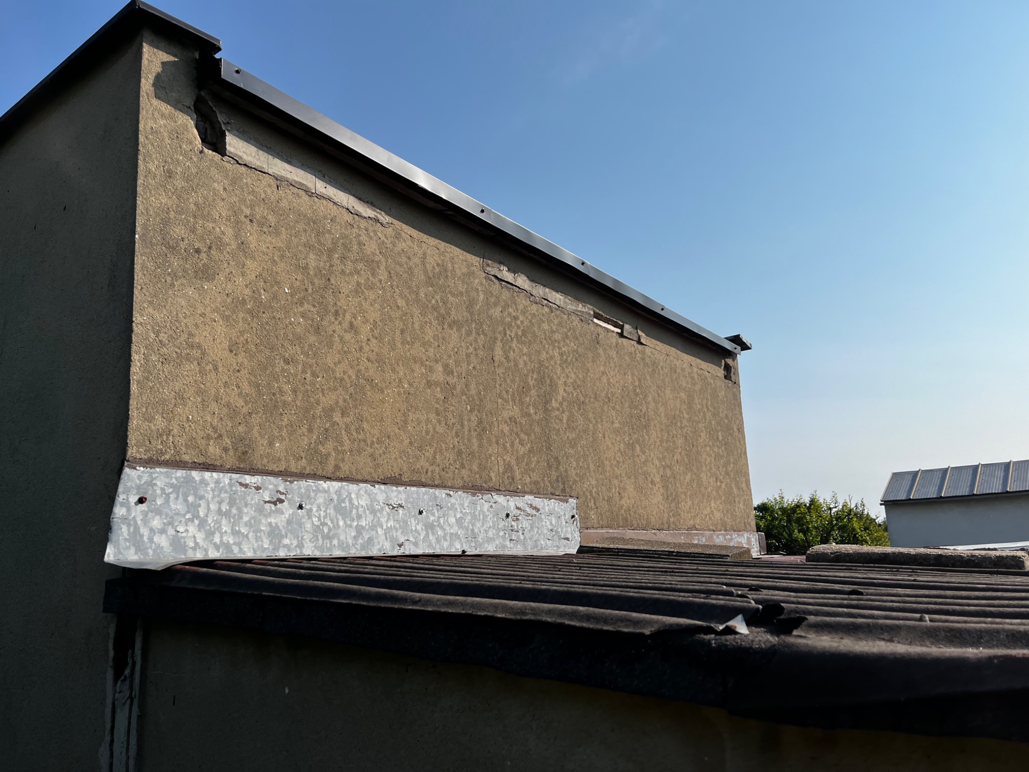 Fragment murowanego budynku z widocznymi uszkodzeniami ściany i dachu z blachy falistej, w tle błękitne niebo.