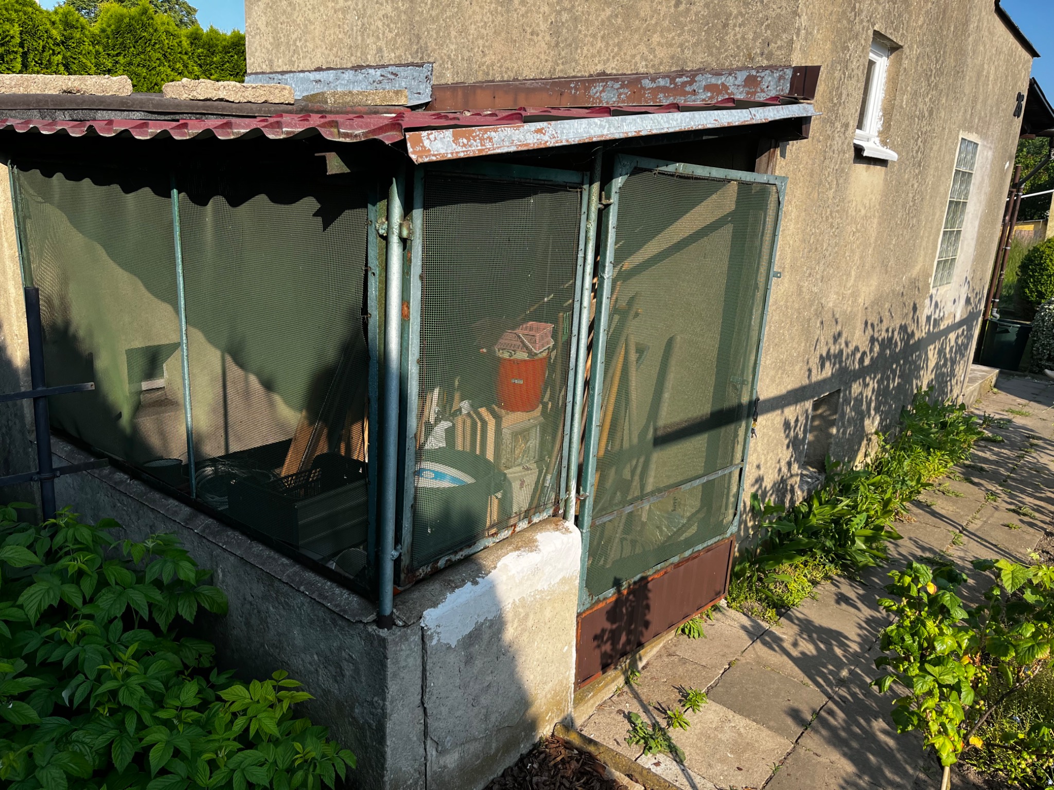 Niewielka, zadaszona komórka gospodarcza z metalową siatką zamiast ścian, przylegająca do budynku z oknami, otoczona zielenią.