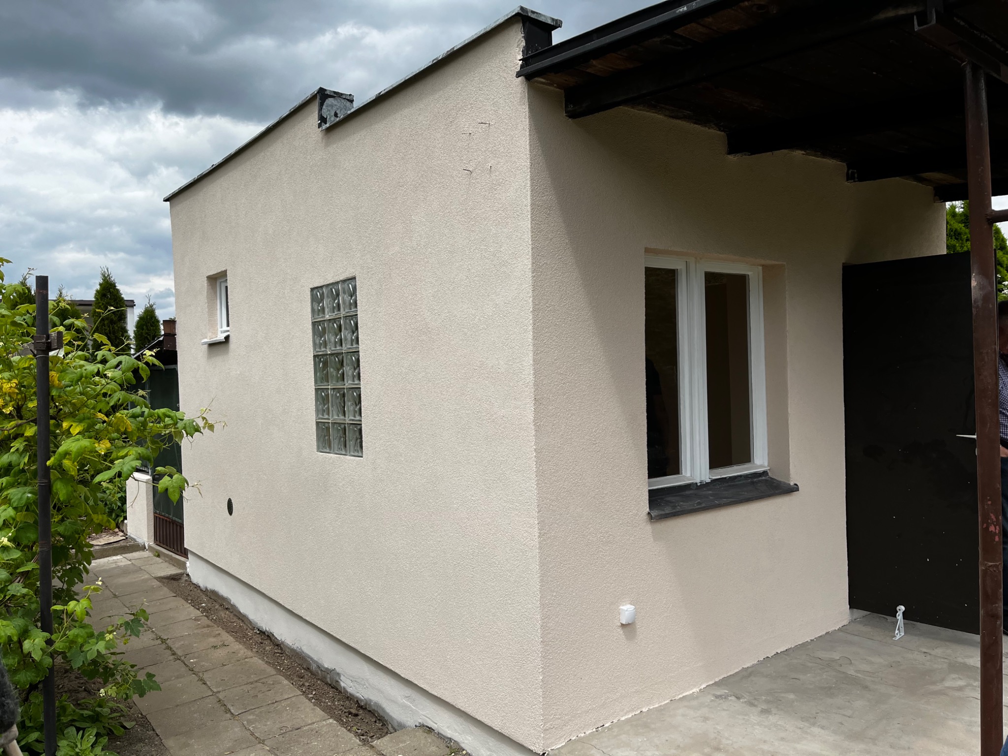 Niewielki, otynkowany na beżowo budynek gospodarczy z oknem z luksferów i drewnianą wiatą, widoczny z zewnątrz w pochmurny dzień.