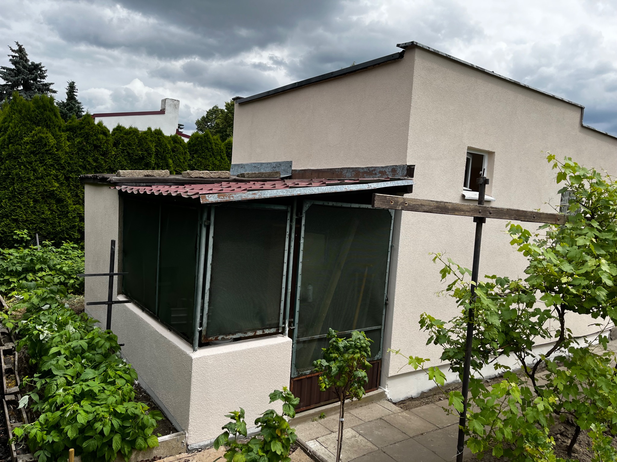 Nieduży, parterowy dom z dobudowaną werandą z metalowymi ramami okiennymi, widok z boku na tle zachmurzonego nieba i zieleni.