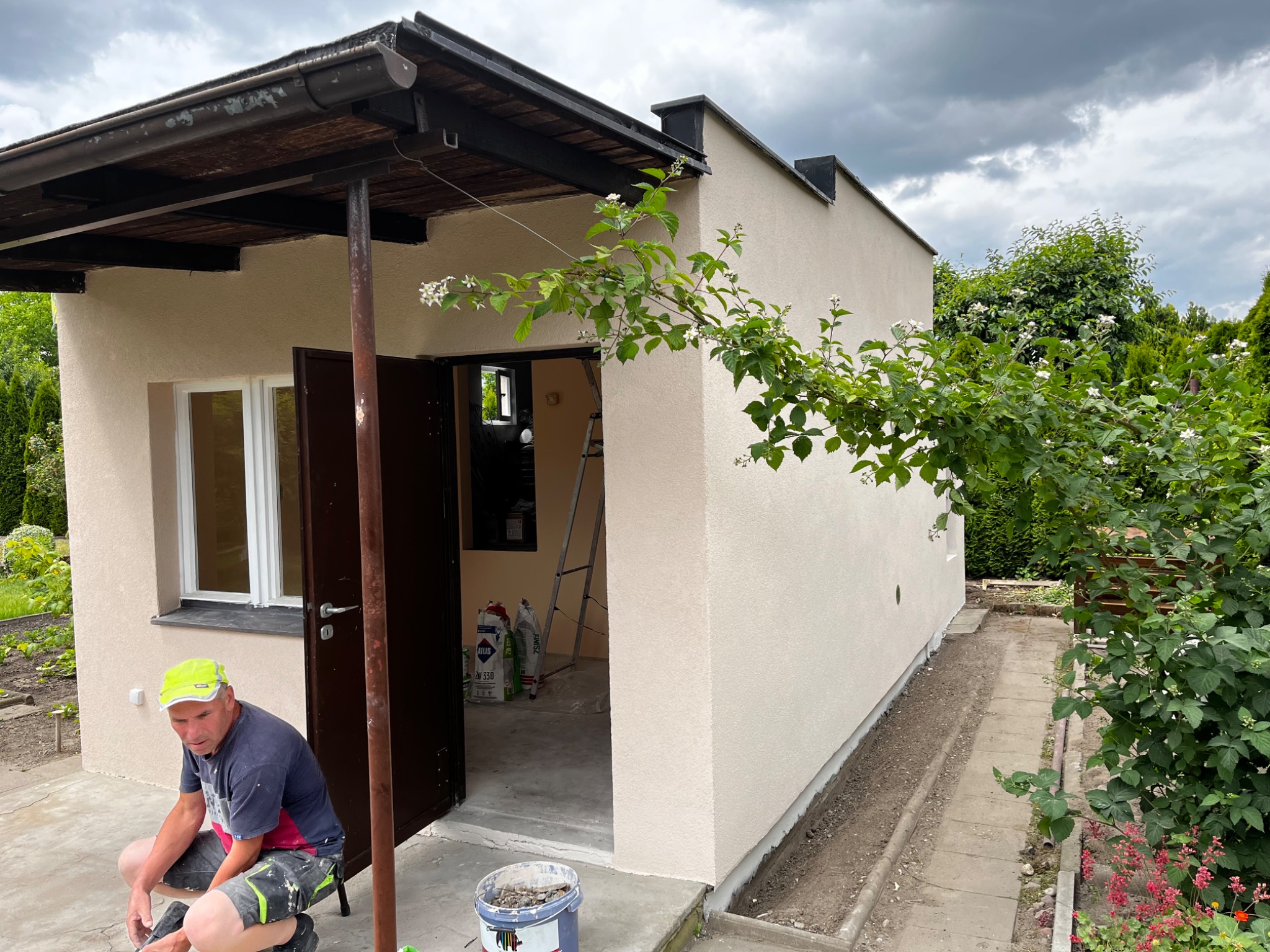 Tynkowana elewacja budynku gospodarczego z widocznym pracownikiem w żółtej czapce, workami z zaprawą wewnątrz i kwitnącą gałęzią na pierwszym planie.