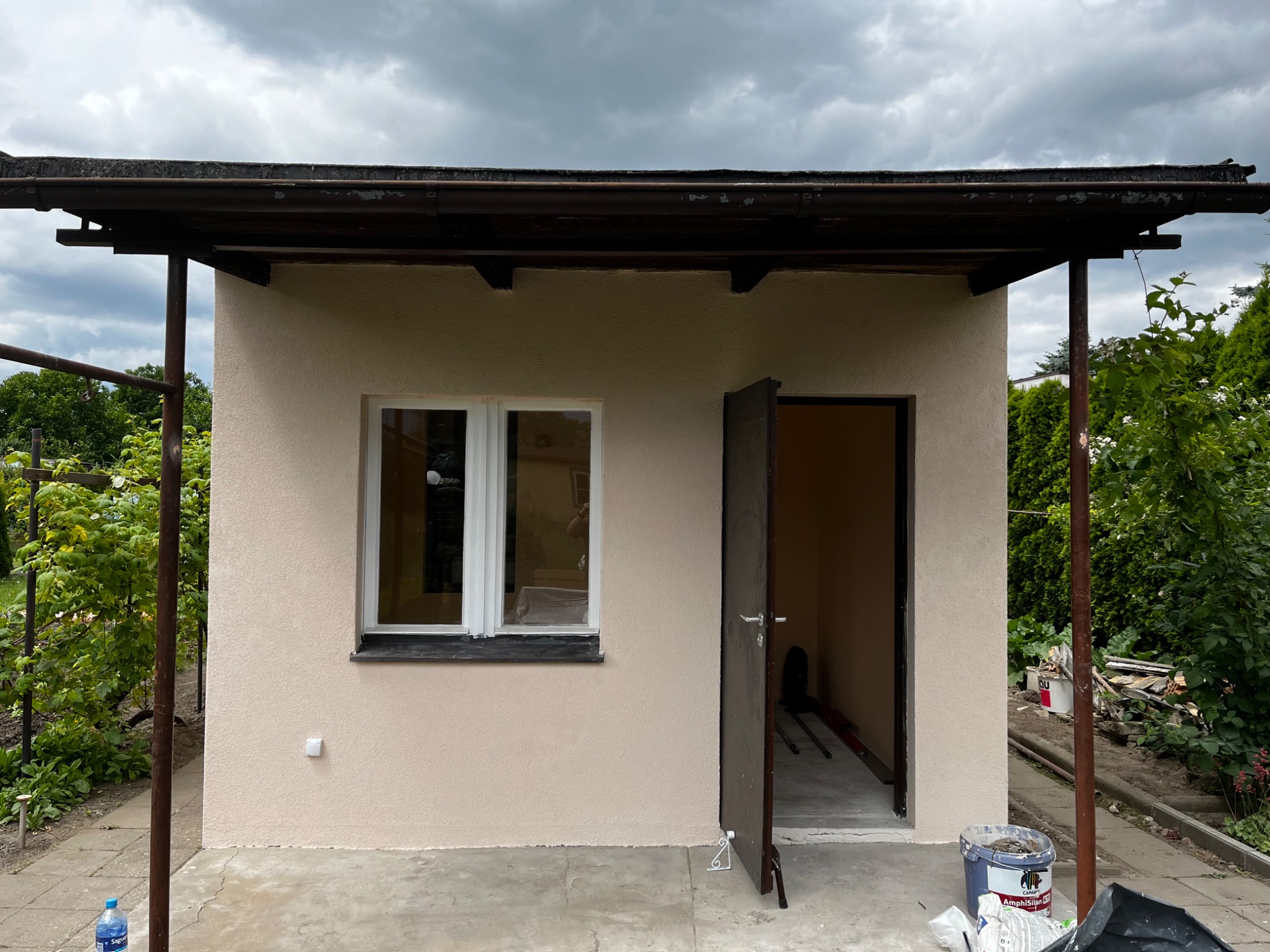 Niewielki budynek gospodarczy z oknem i otwartymi drzwiami w Poznaniu, z wykończoną elewacją w odcieniach beżu, widoczny podczas prac wykończeniowych na zewnątrz, z fragmentami materiałów...