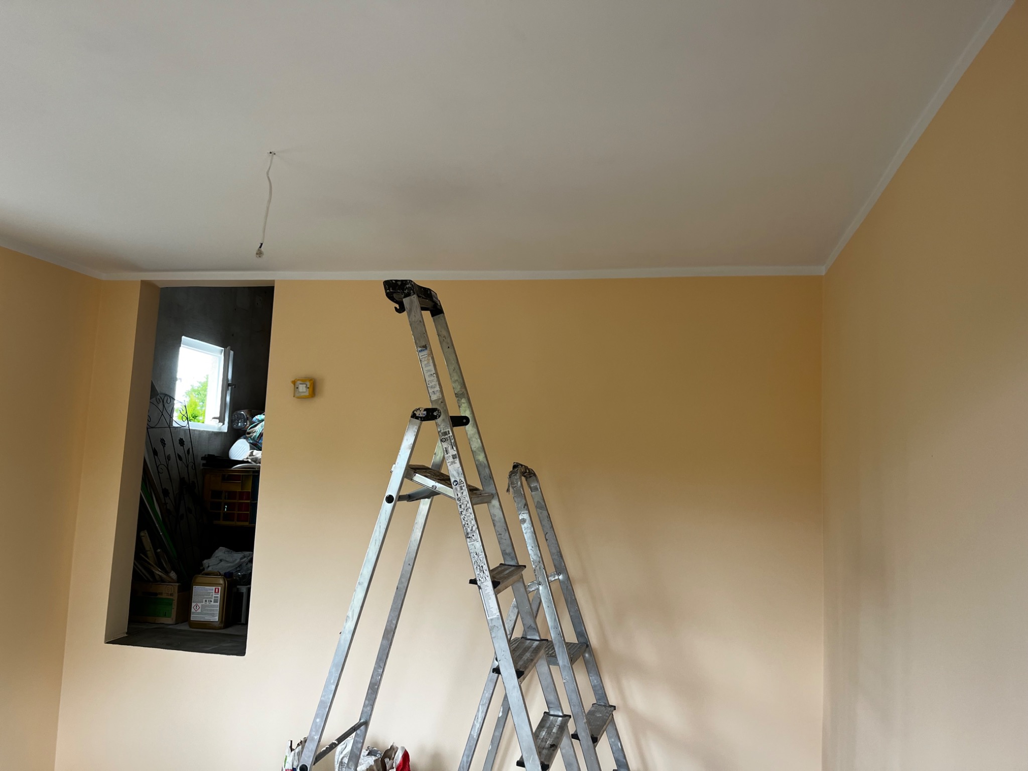 Wnętrze w trakcie malowania: beżowe ściany z białym wykończeniem pod sufitem, aluminiowa drabina rozstawiona w pokoju, widok na zagracone pomieszczenie przez otwór w ścianie.