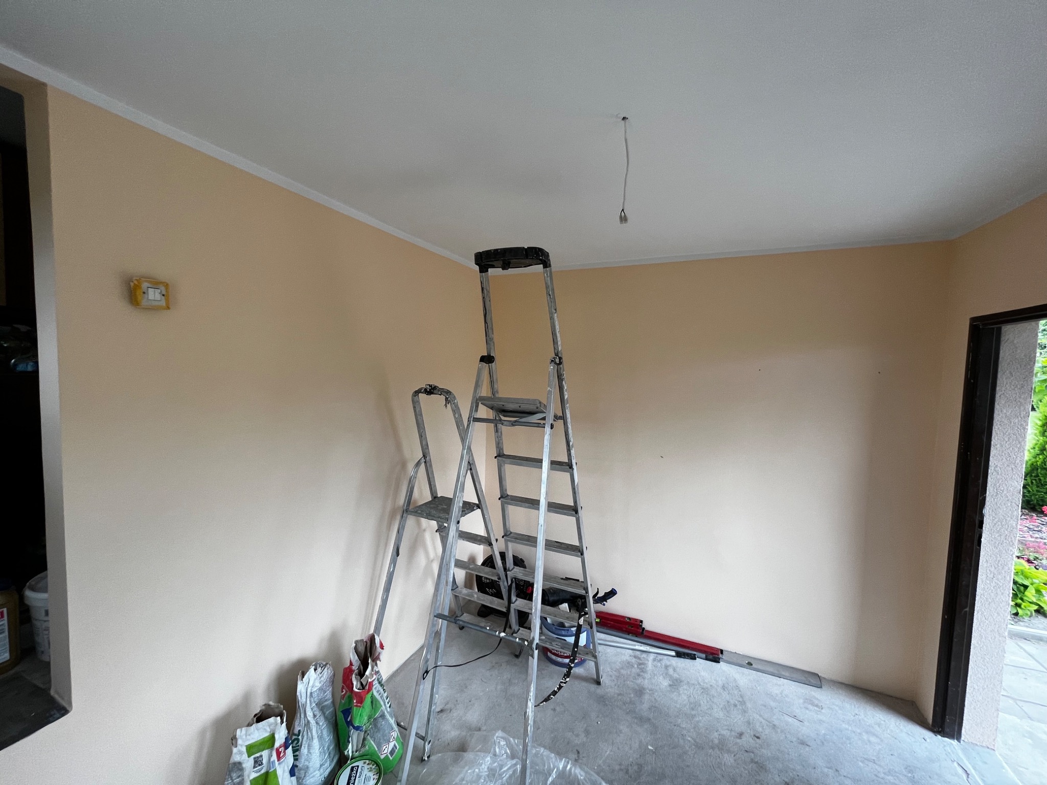 Pomieszczenie w trakcie malowania ścian na jasny beż, widoczne dwie drabiny, worki z zaprawą, narzędzia malarskie i wystający kabel z sufitu.