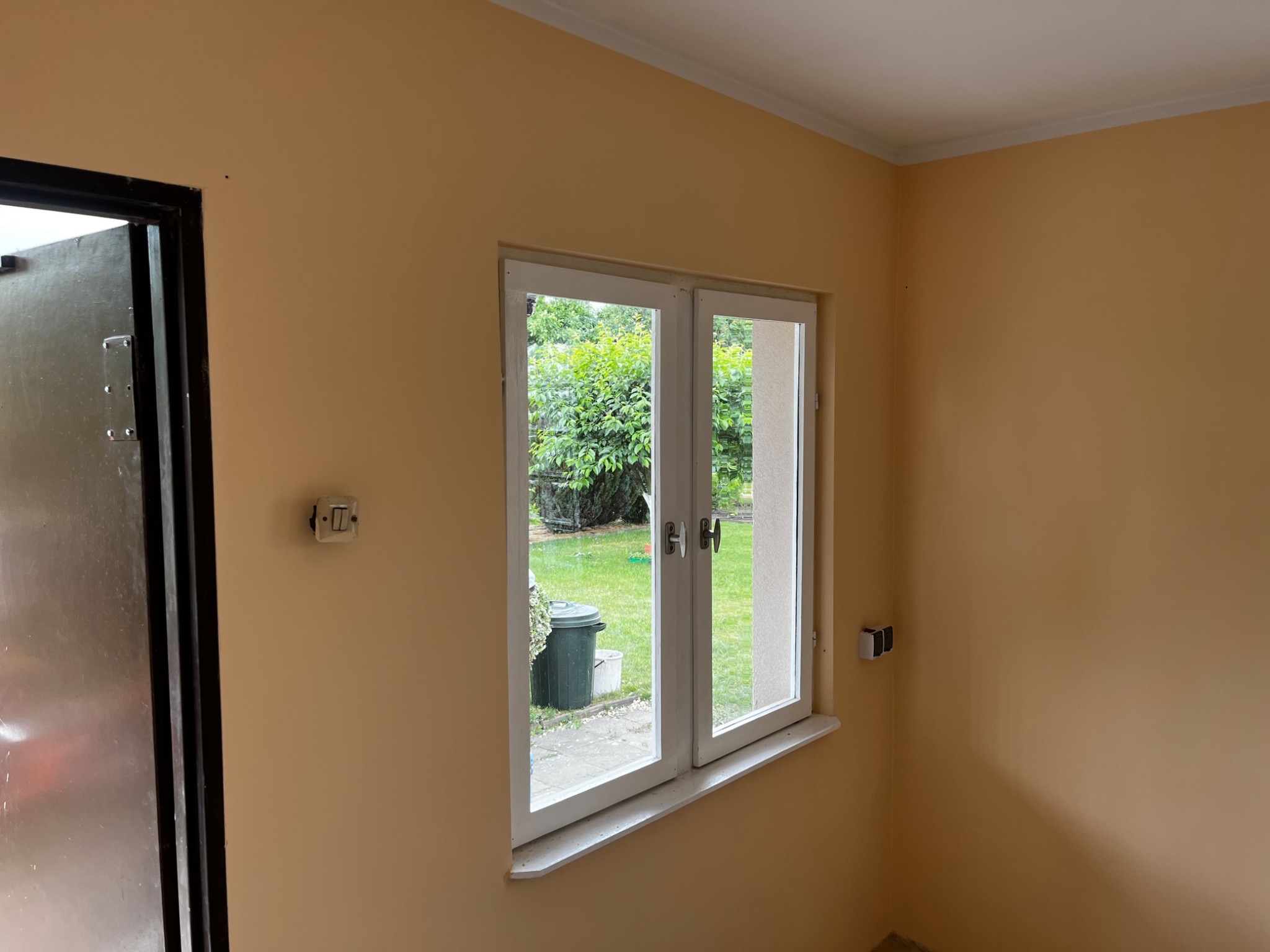 Wnętrze pokoju w trakcie remontu z beżowymi ścianami, białym oknem z widokiem na ogród i czarnymi drzwiami po lewej stronie.