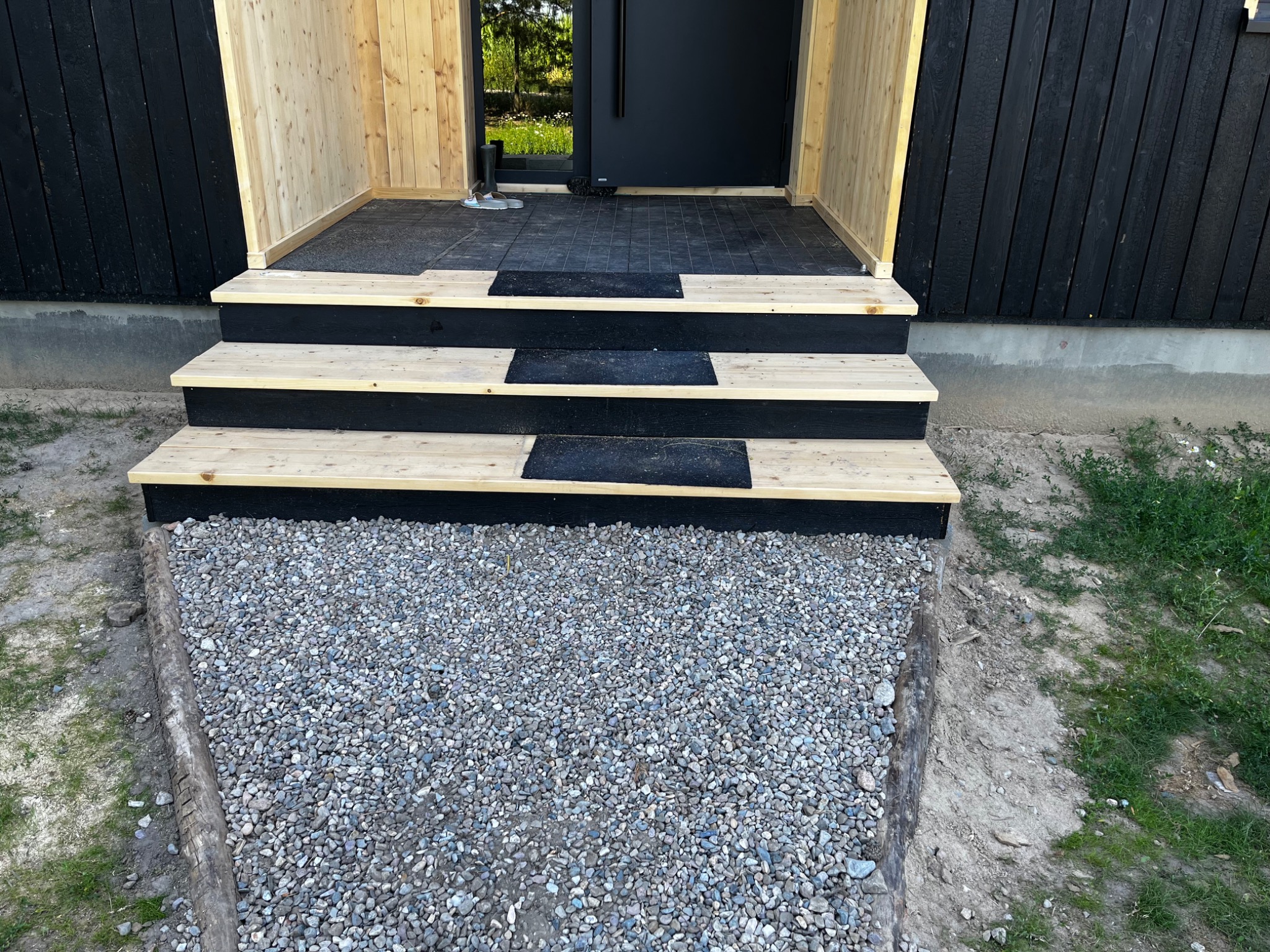 Nowoczesne wejście do domu z czarną elewacją i drewnianymi schodami, wykończone żwirem.