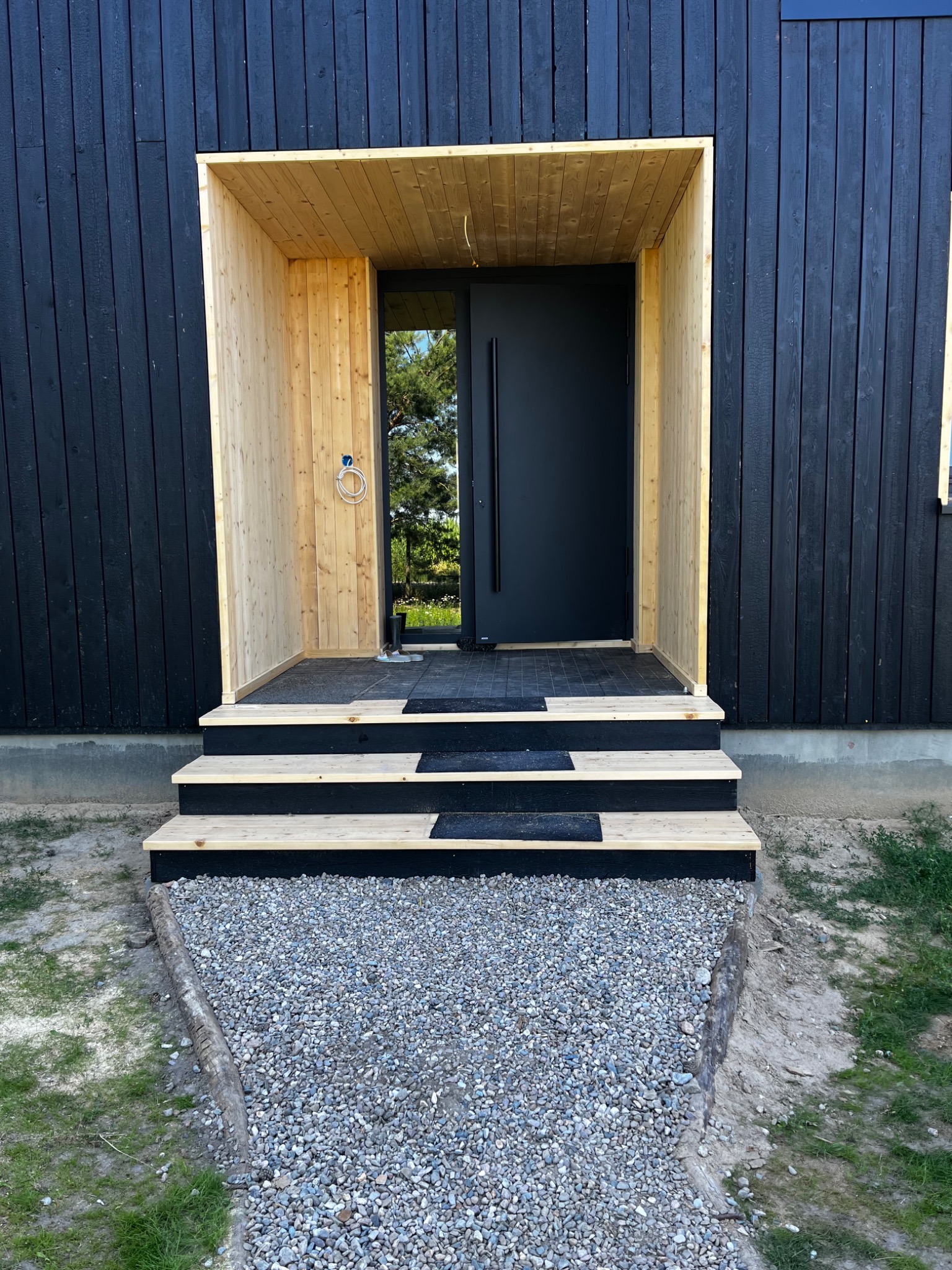 Nowoczesny dom z czarną drewnianą elewacją, jasnym drewnianym gankiem i minimalistycznymi schodami wejściowymi prowadzącymi do czarnych drzwi.