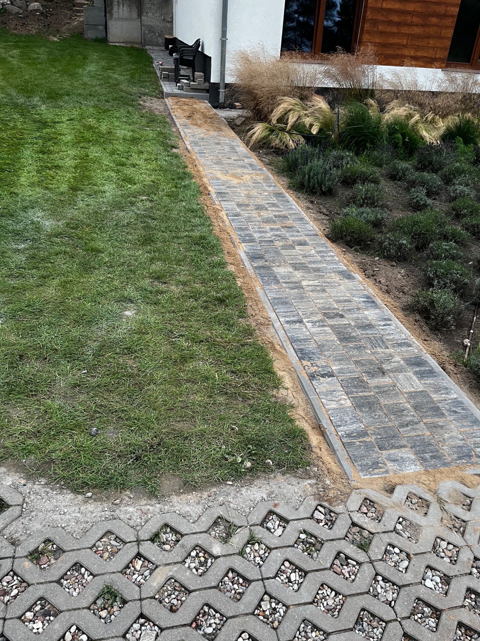 Ułożona ścieżka z kostki brukowej prowadząca do domu, obok trawnik i rabaty z roślinami ozdobnymi, widoczne elementy ażurowych płyt betonowych.
