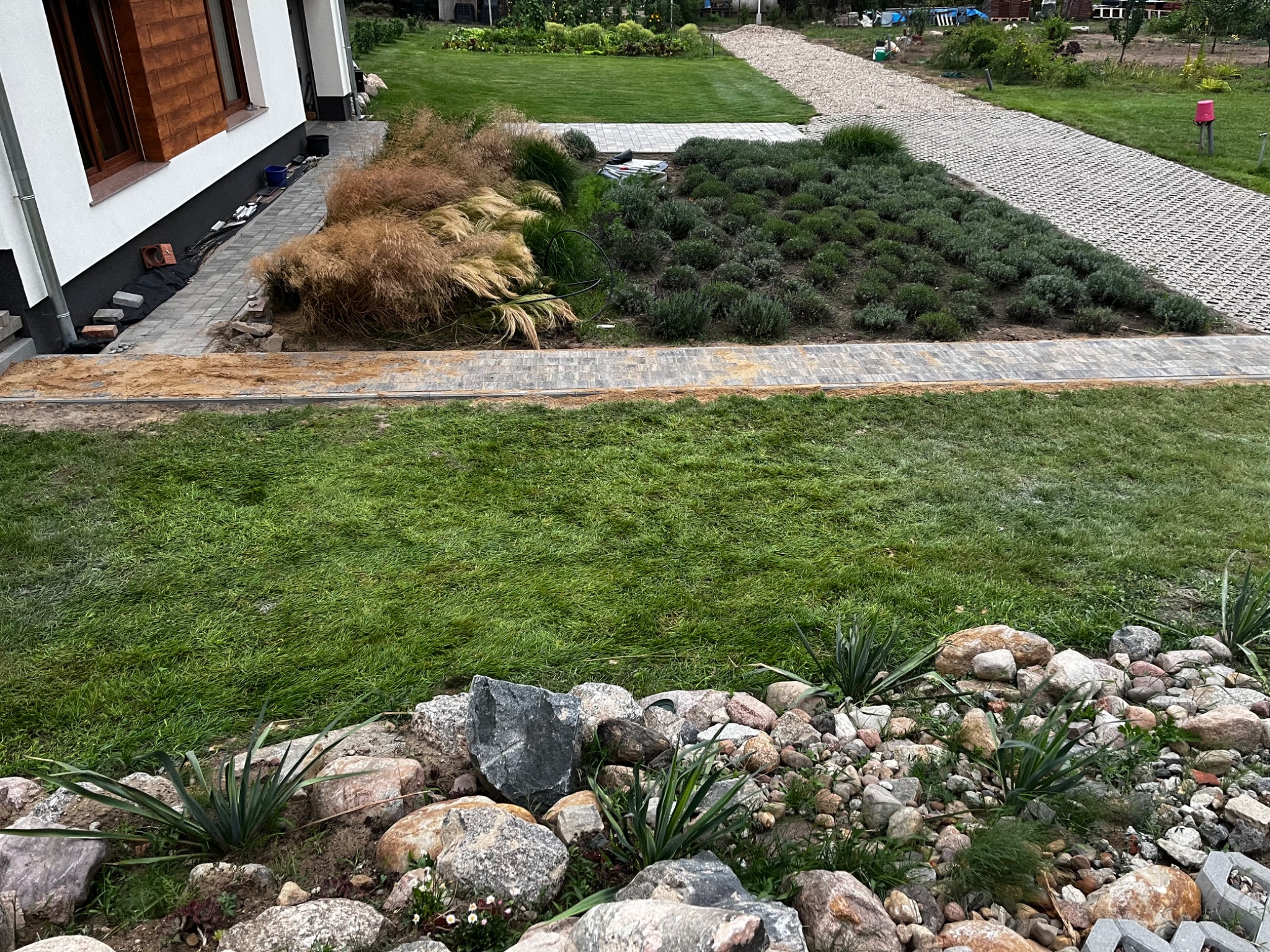 Fragment ogrodu z nowo założonymi rabatami, ścieżką z ażurowych płyt i kamiennym obrzeżem, widoczne trawy ozdobne i niskie krzewy.