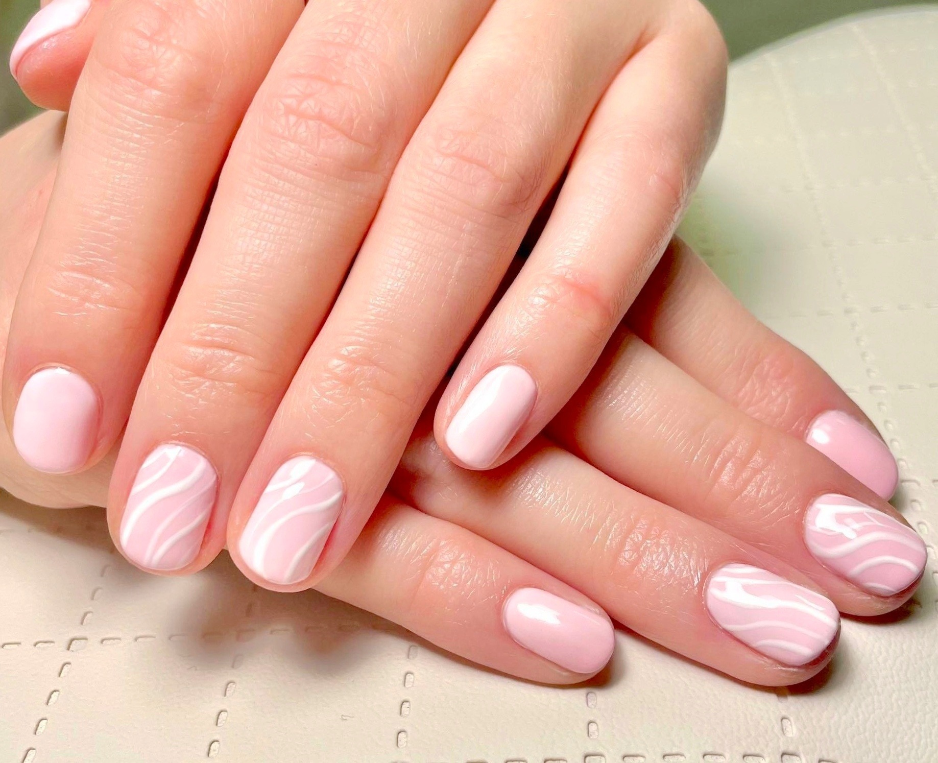 Dłonie z pastelowym różowym manicure i białymi marmurkowymi wzorami na niektórych paznokciach, ułożone na jasnym tle.