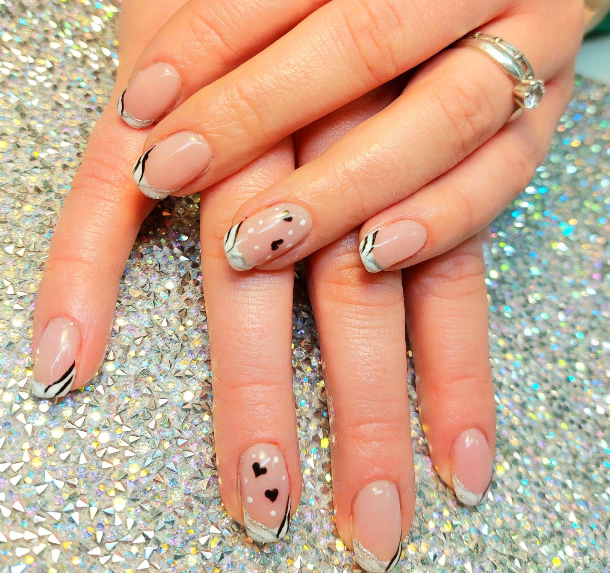 Dłonie z manicure hybrydowym: jasnoróżowe paznokcie z białymi końcówkami zdobionymi czarnymi paskami i serduszkami, na błyszczącym srebrnym tle, pierścionek na palcu.