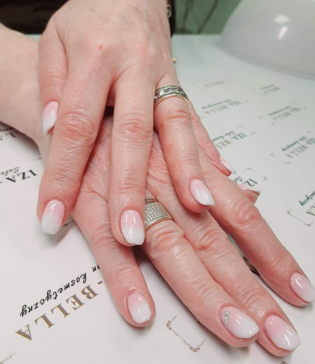 Dłonie kobiety z delikatnym, ombre zdobieniem paznokci w odcieniach różu i bieli, ozdobione srebrnymi drobinkami na jednym paznokciu, na tle z logo salonu kosmetycznego.