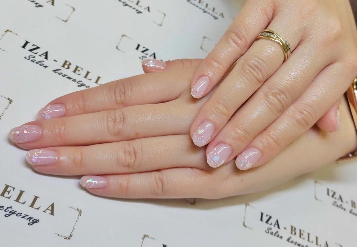 Delikatne, jasnoróżowe paznokcie z subtelnymi, opalizującymi drobinkami brokatu, zdobione na końcach białymi i niebieskimi akcentami, na tle z logo salonu kosmetycznego IZA-BELLA, z widocznym...