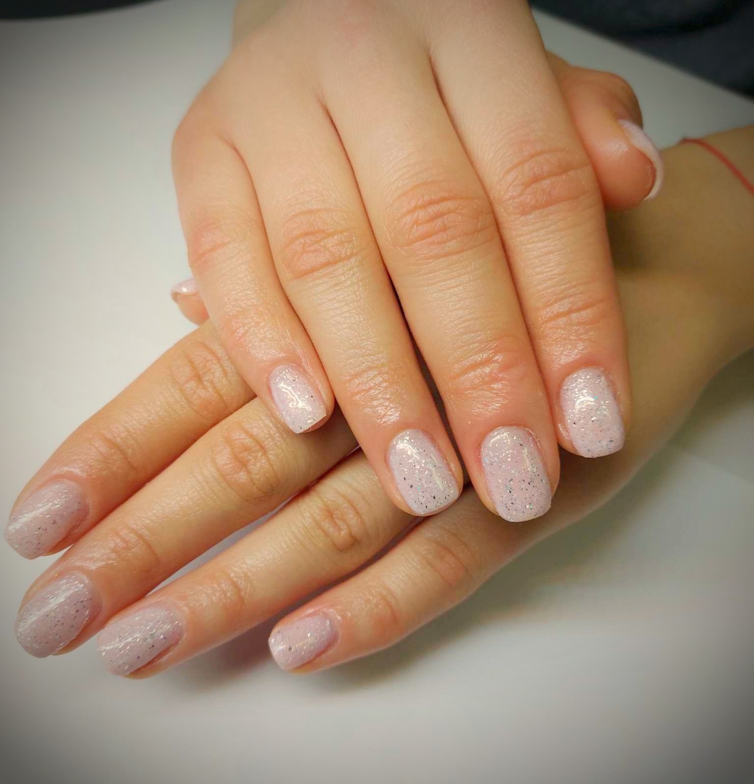 Dłonie kobiety ze świeżo pomalowanymi paznokciami w jasnoróżowym kolorze z delikatnym srebrnym brokatem na tle białego stołu.