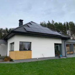 Nowoczesny dom jednorodzinny z panelami słonecznymi na dachu, widok z trawnika, w tle las iglasty i pochmurne niebo.