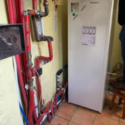 Biała pompa ciepła Daikin w trakcie instalacji, widoczne liczne rury hydrauliczne w kolorze czerwonym i niebieskim, instalator w górnym prawym rogu zdjęcia, instrukcja montażu na obudowie urządzenia.