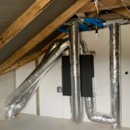 Instalacja systemu wentylacji mechanicznej z odzyskiem ciepła w surowym pomieszczeniu poddasza: widoczne srebrne, izolowane rury wentylacyjne, czarna centrala wentylacyjna, niebieskie przewody...