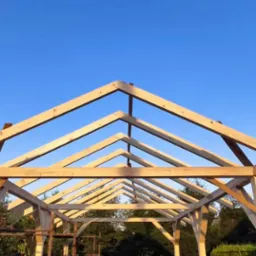 Konstrukcja drewniana z belek pod przyszłą pergolę lub altanę, widok z dołu na niebieskie niebo, w tle zieleń drzew i krzewów.