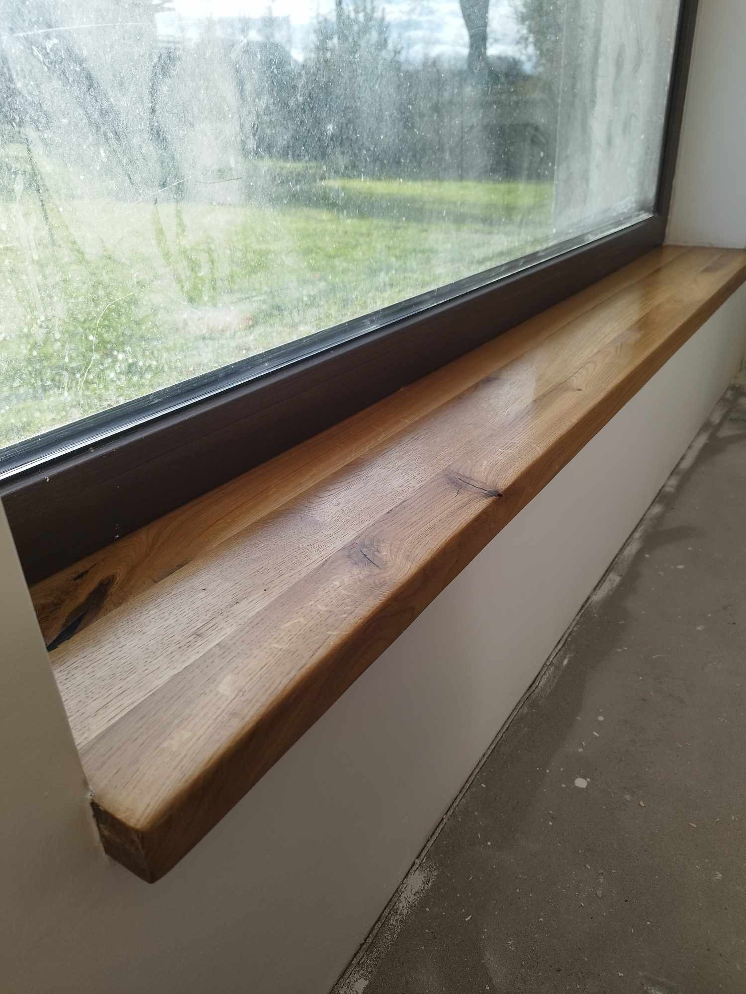 Nowy, lakierowany, drewniany parapet z widocznym usłojeniem, zamontowany pod oknem z widokiem na zewnątrz. Ściana pomalowana na biało, podłoga betonowa.