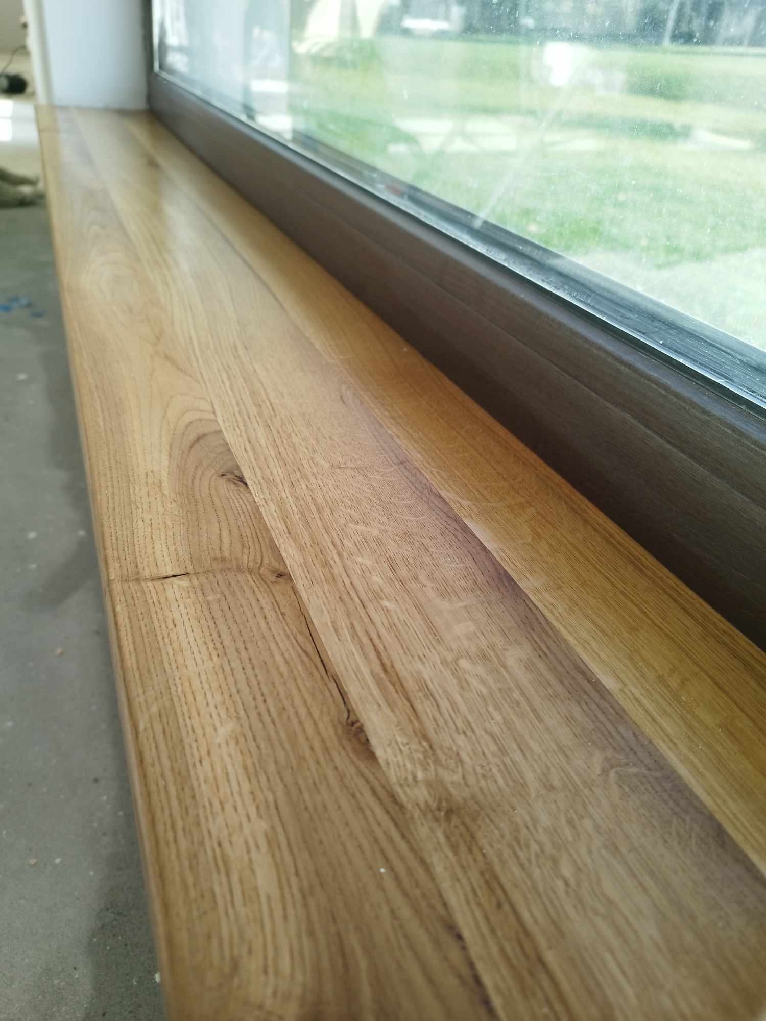 Jasny, dębowy parapet z widocznym usłojeniem i naturalnymi pęknięciami, zamontowany pod oknem z widokiem na zieleń.