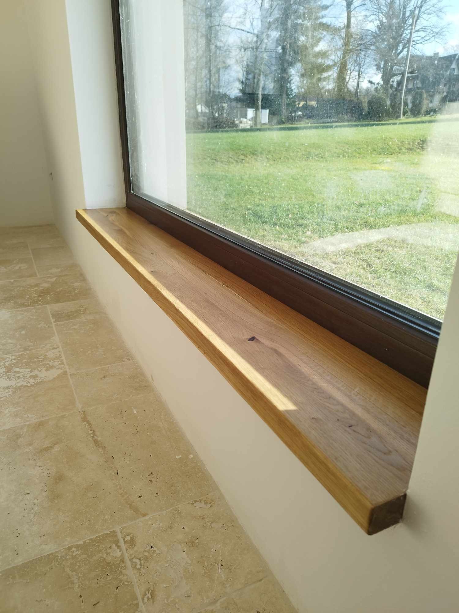 Jasny, dębowy parapet wewnętrzny z widocznym usłojeniem, zamontowany w oknie z ciemną ramą, z widokiem na trawnik i drzewa.