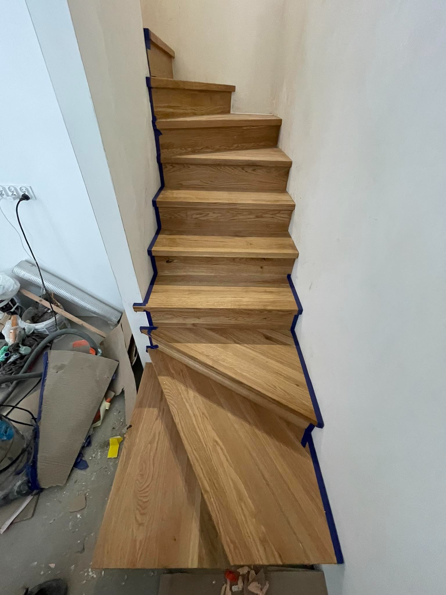 Drewniane schody w trakcie montażu, zabezpieczone niebieską taśmą malarską, widoczne ślady prac remontowych i narzędzia na podłodze.