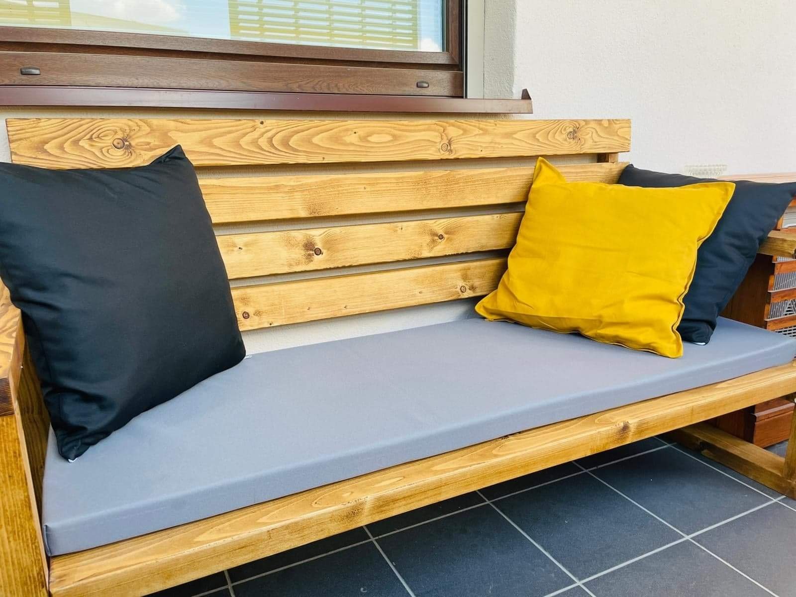 Drewniana ławka ogrodowa z szarą poduszką i czarno-żółtymi poduszkami dekoracyjnymi, wykonana w Krakowie, ustawiona na ciemnej, kafelkowej podłodze balkonu przy białej ścianie z brązowym oknem.