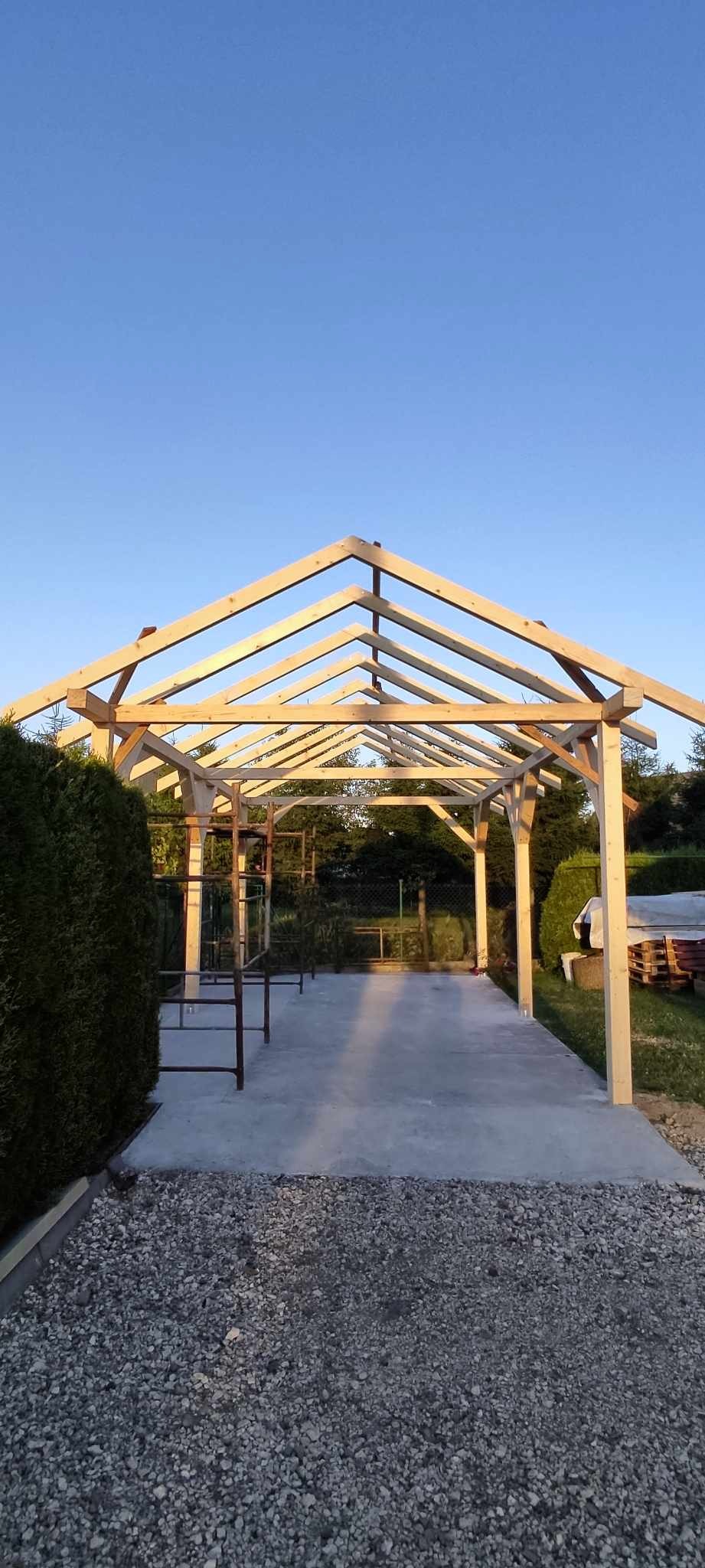 Konstrukcja drewniana zadaszenia w trakcie budowy, widoczne słupy, krokwie, betonowa wylewka, krzewy w tle, wczesny wieczór.