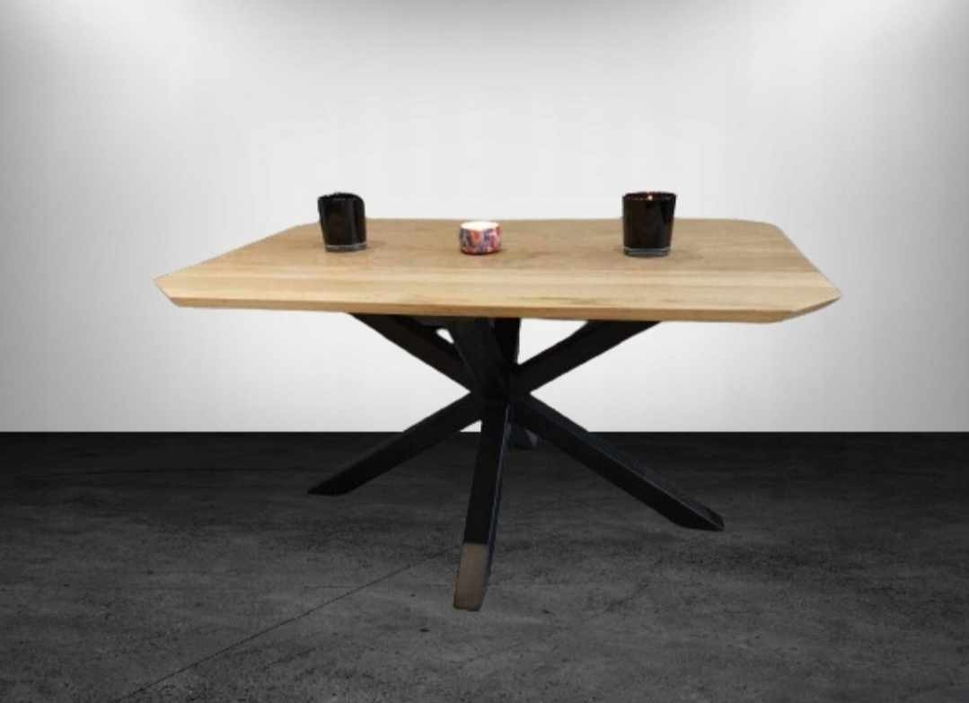 Kwadratowy stół z blatem z jasnego drewna i czarną, geometryczną podstawą, na blacie dwie czarne świeczki i jedna ozdobna.