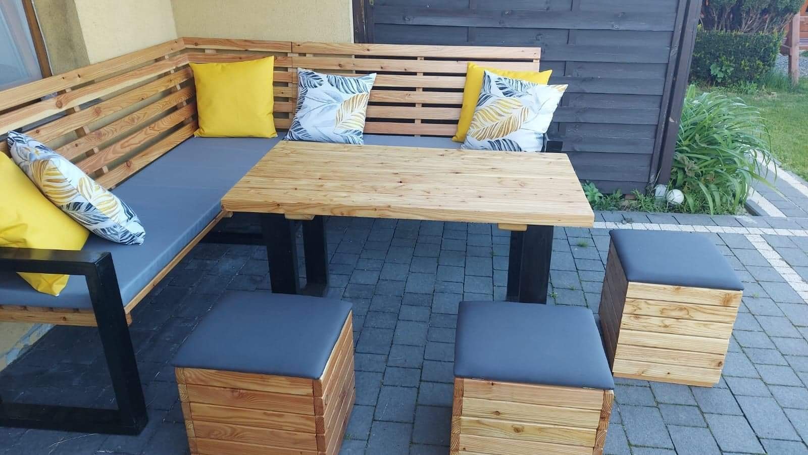 Drewniany narożnik z poduszkami, stół z czarnymi nogami i pufy z szarymi siedziskami na brukowanym tarasie.