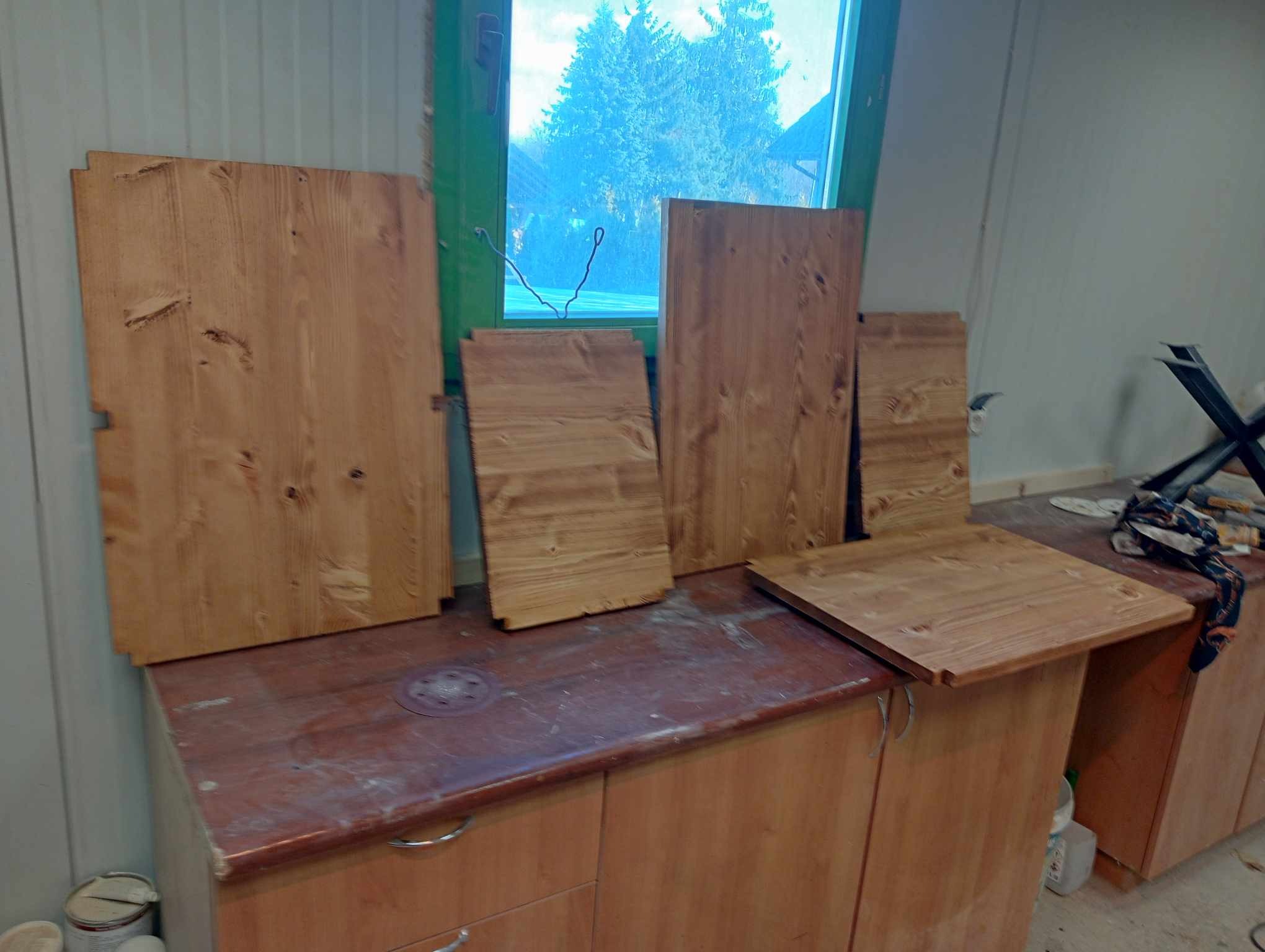 Surowe, drewniane elementy szafy wnękowej, częściowo obrobione, ustawione na blacie roboczym w warsztacie, widoczne słoje drewna i niedoskonałości, okno w tle.