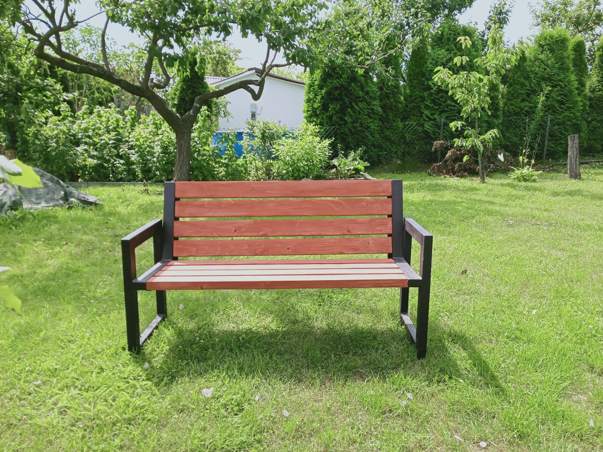 Drewniana ławka ogrodowa z oparciem w kolorze brązowym i czarną ramą, ustawiona na trawniku w ogrodzie z drzewami i krzewami w tle.