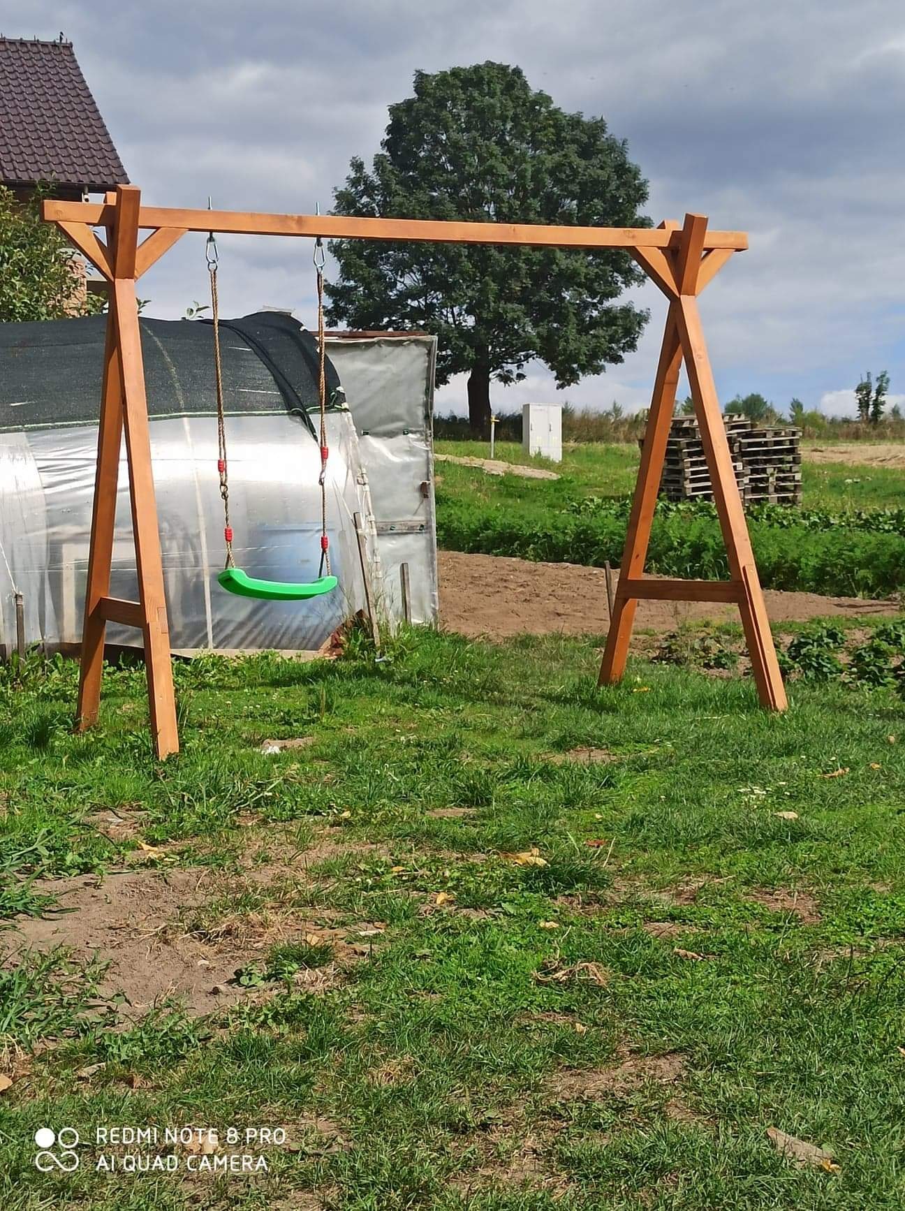 Drewniana huśtawka ogrodowa z zielonym siedziskiem, zawieszona na sznurach, na tle ogrodu z widoczną szklarnią i stertą palet.