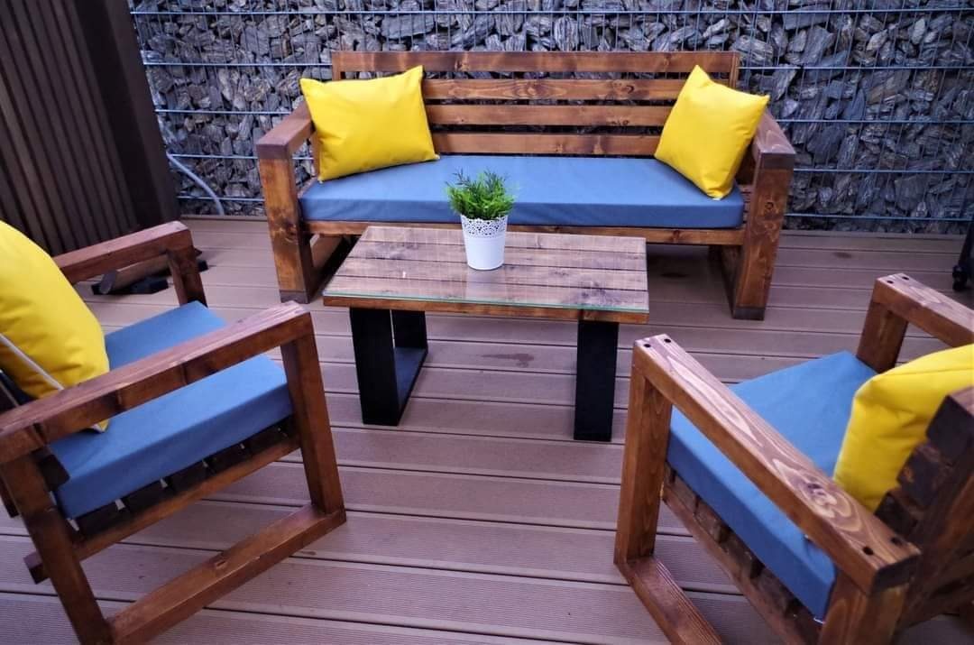 Zestaw mebli ogrodowych z ciemnego drewna: ławka z żółtymi poduszkami, dwa fotele z niebieskimi poduszkami i stolik kawowy z blatem z desek i szklanym obramowaniem, na drewnianym tarasie, w tle...