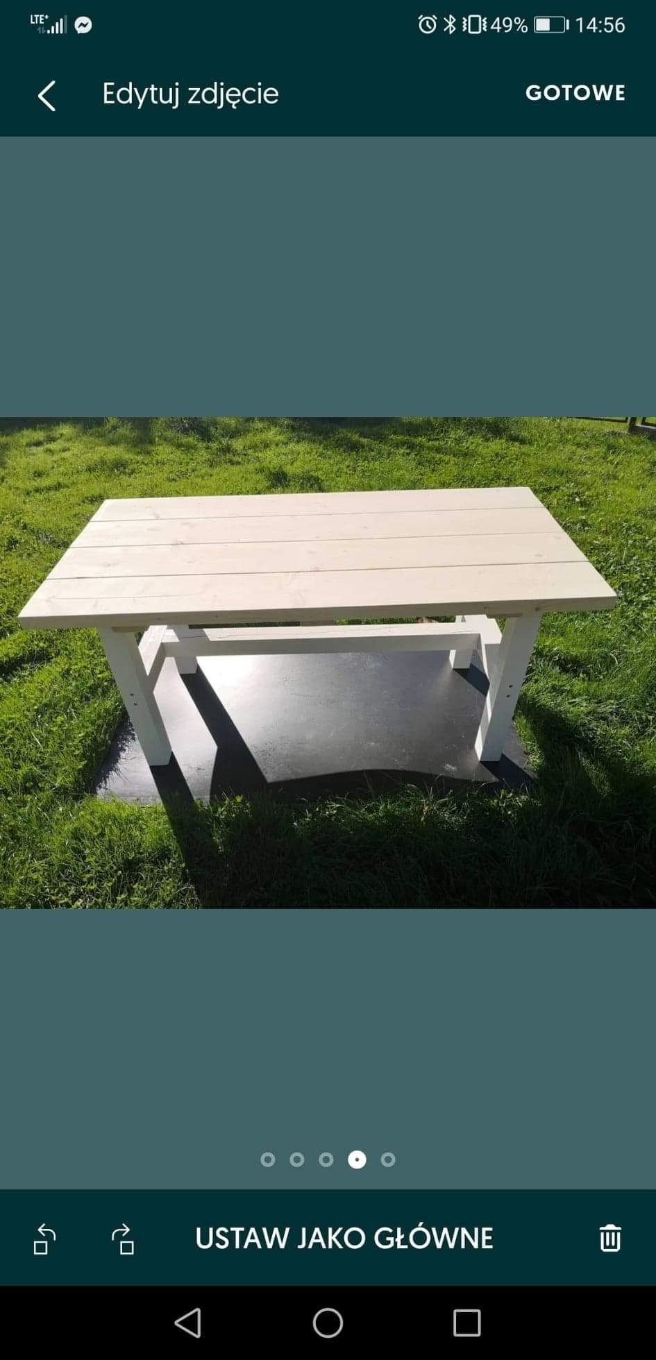 Stół ogrodowy z jasnego drewna, ustawiony na czarnej macie na trawie, widoczne cienie, ujęcie z lekkiej góry.